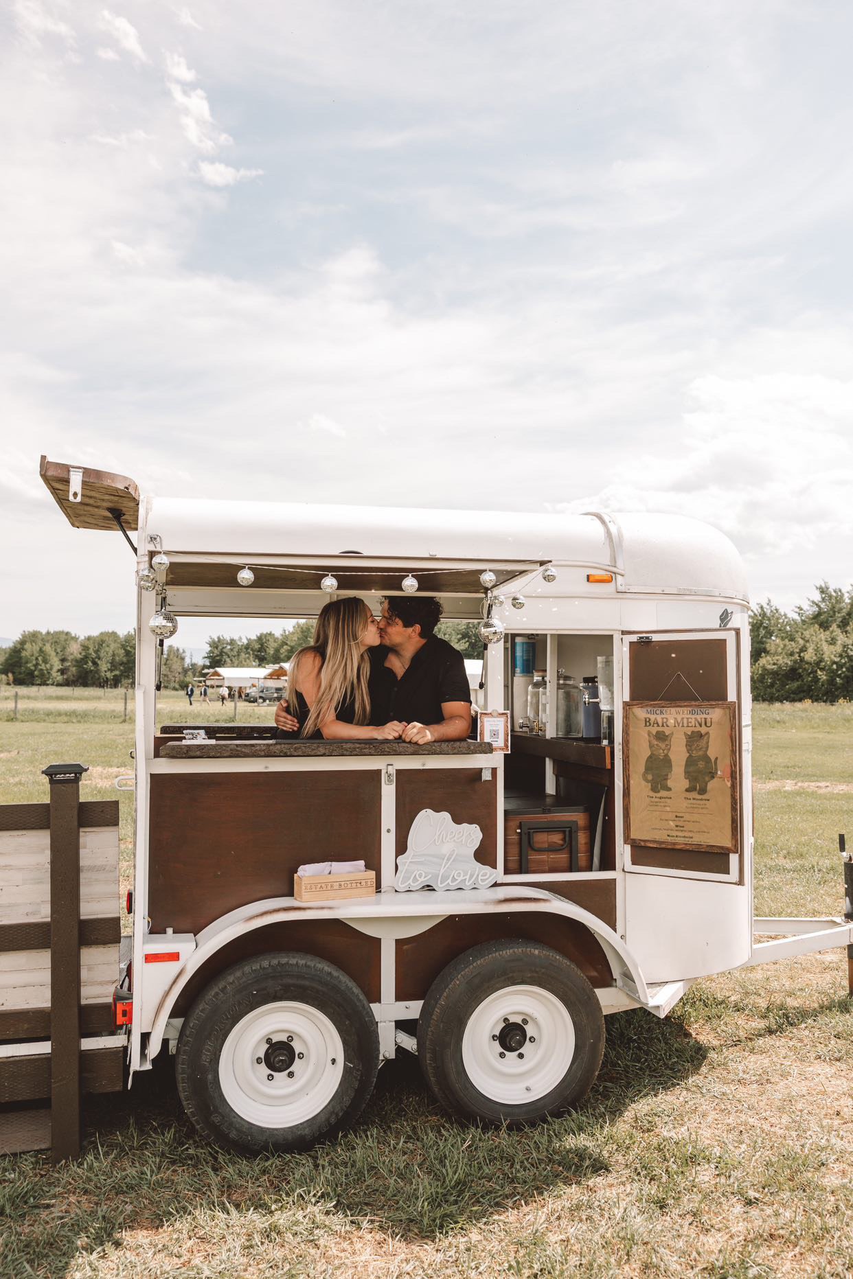 Owners of My Pour Decisions Mobile Bar in Montana,  horse trailer mobile bar and decorative signs, under a cloudy sky.