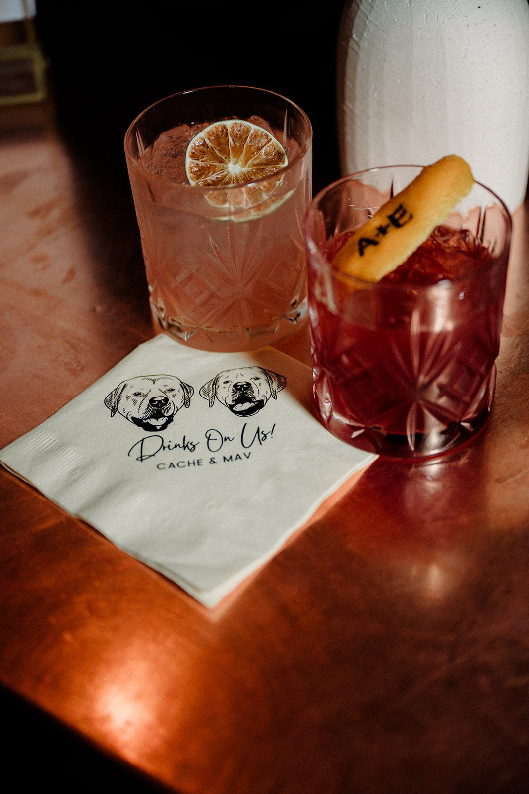 Two cocktails with a napkin underneath that has a dog illustration and the text 'Drinks On Us!" Branded Orange Peel
