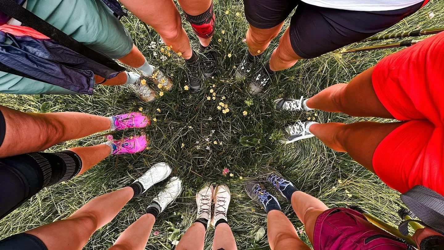 A group of people with various colored shorts and shoes standing in a circle on grassy ground with small yellow and pink flowers.