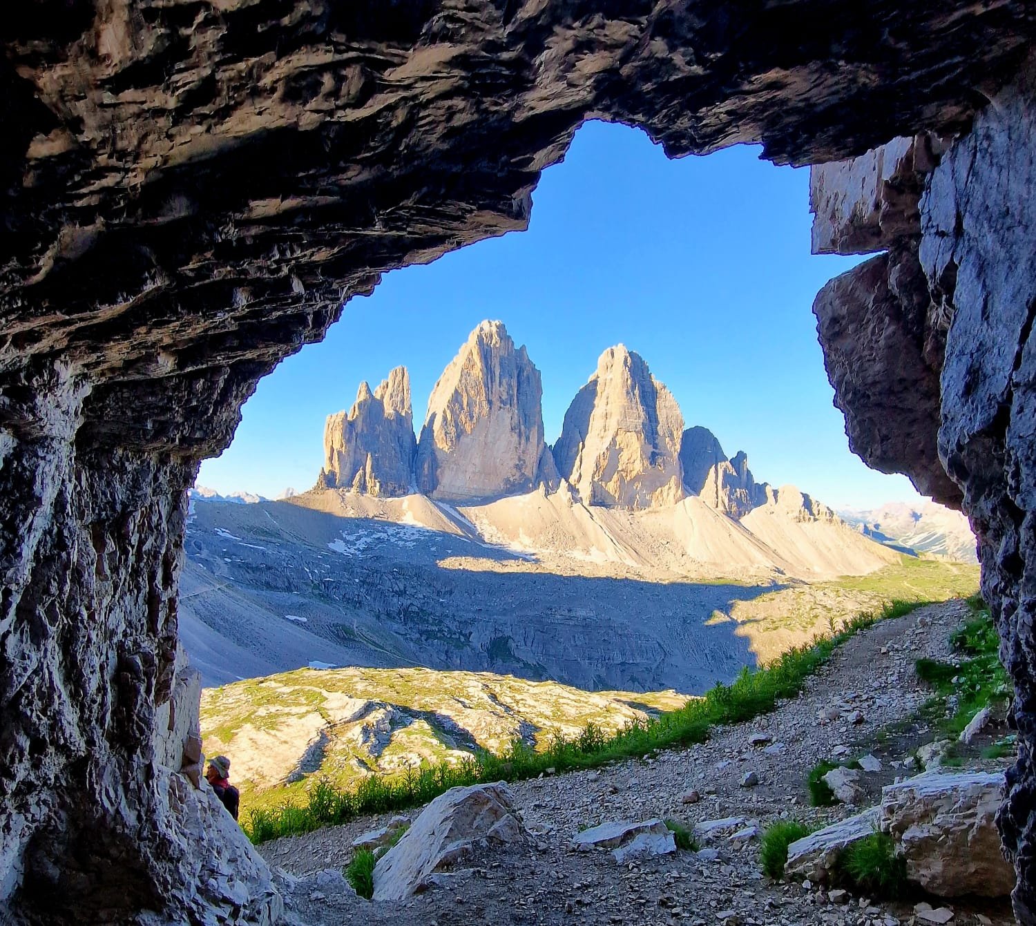 View of Tre Cime through a rocky cave opening with a hiking trail and green grass in the foreground under a clear blue sky.
