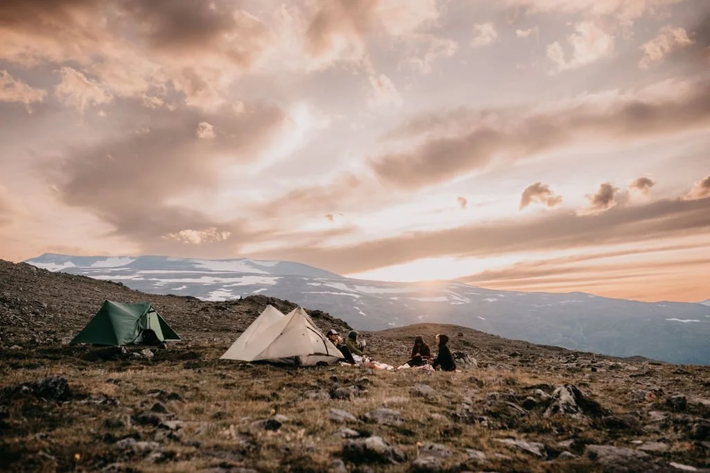 Two tents set up on a rocky, grassy hillside during sunset with two people sitting nearby, overlooking mountains with patches of snow under a partly cloudy sky.
