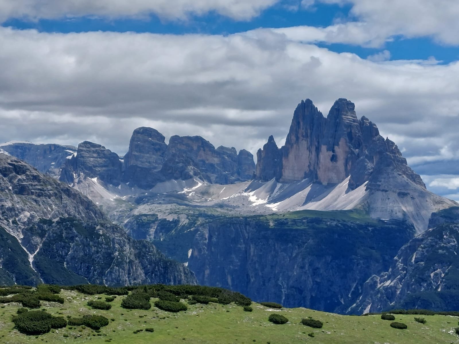 Scenic mountain landscape with rocky peaks, some snow patches, a green meadow with bushes in the foreground, and partly cloudy sky.