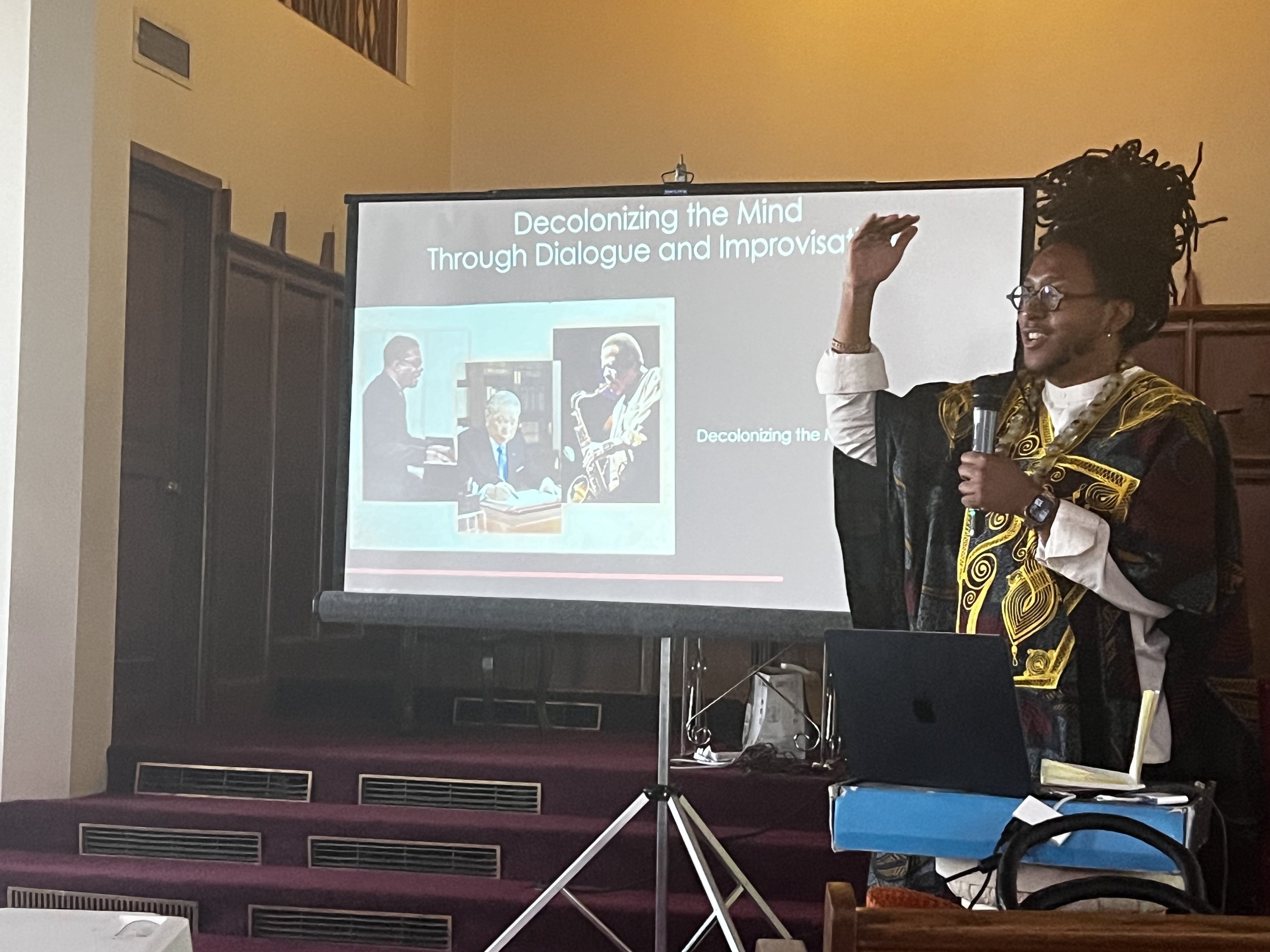 Loki Karuna stands beside a projector screen with the title "Decolonizing the Mind through Dialogue and Improvisation, speaking into a mic held in one hand. The other hand is raised.