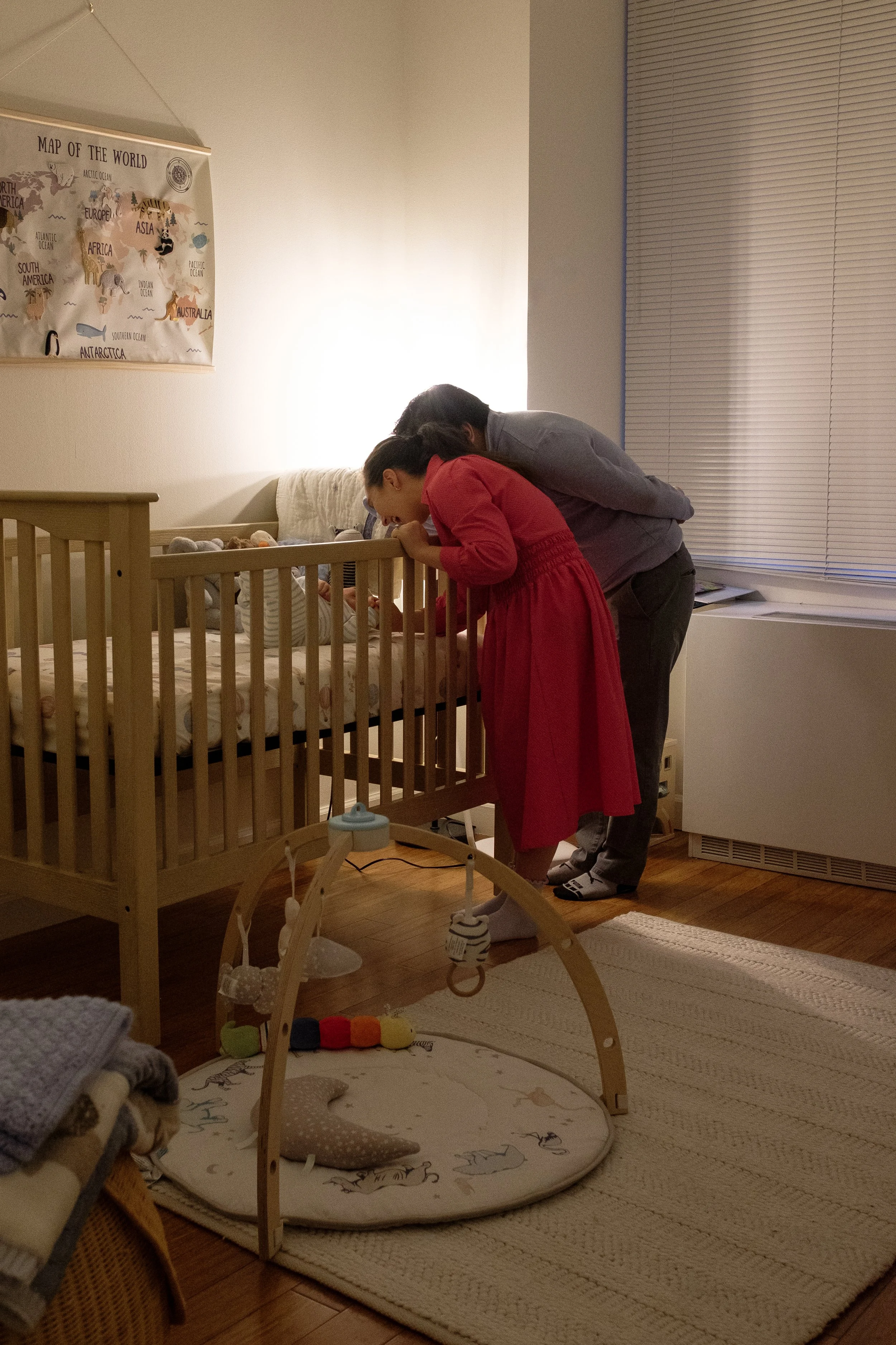 A couple looking into a baby crib in a nursery.  There's a baby laying on the crib, and a colorful activity gym on the floor nearby. The room has a world map poster on the wall and a window with blinds.