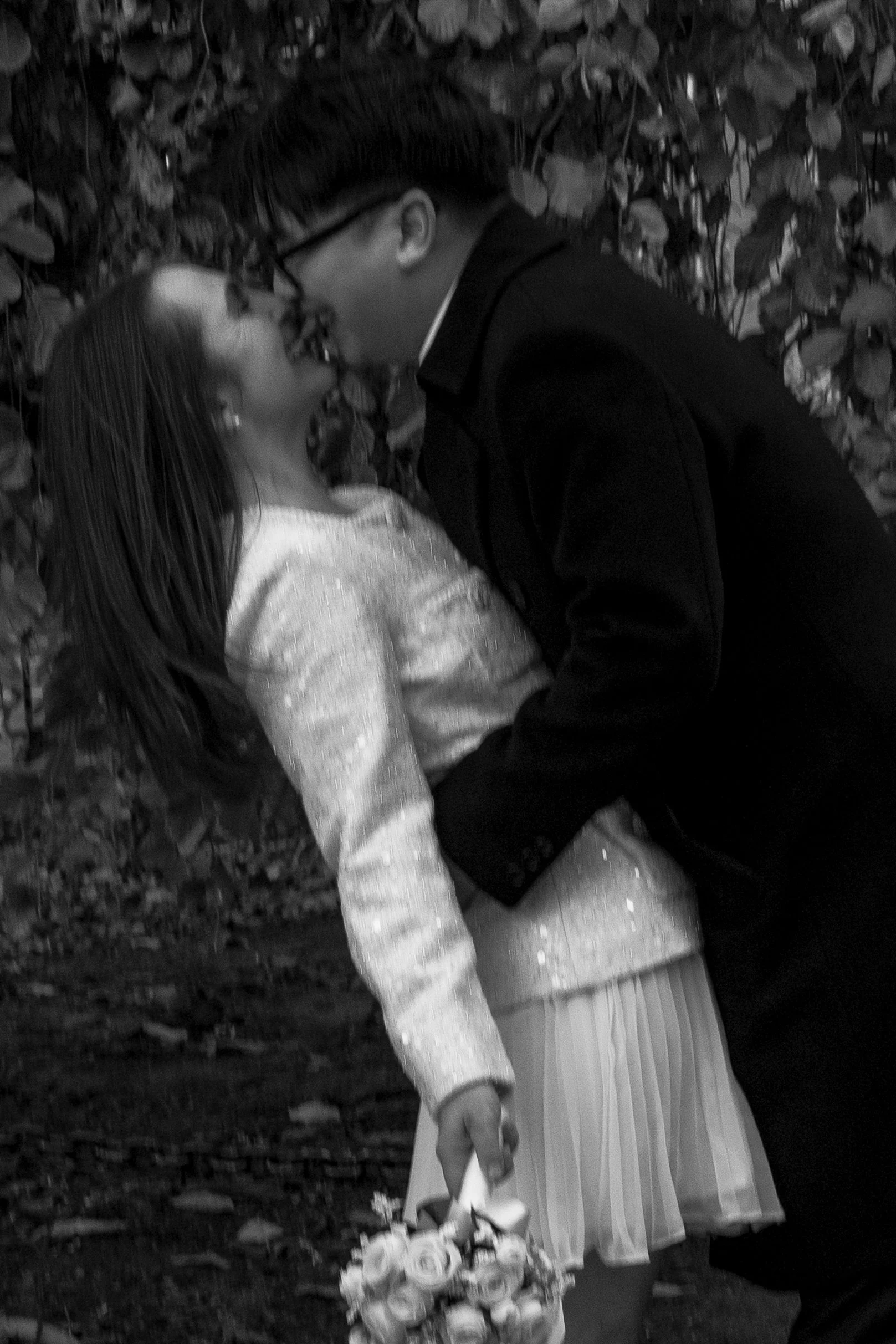 A black-and-white photo of a couple sharing a kiss outdoors, with the woman holding a bouquet of flowers.