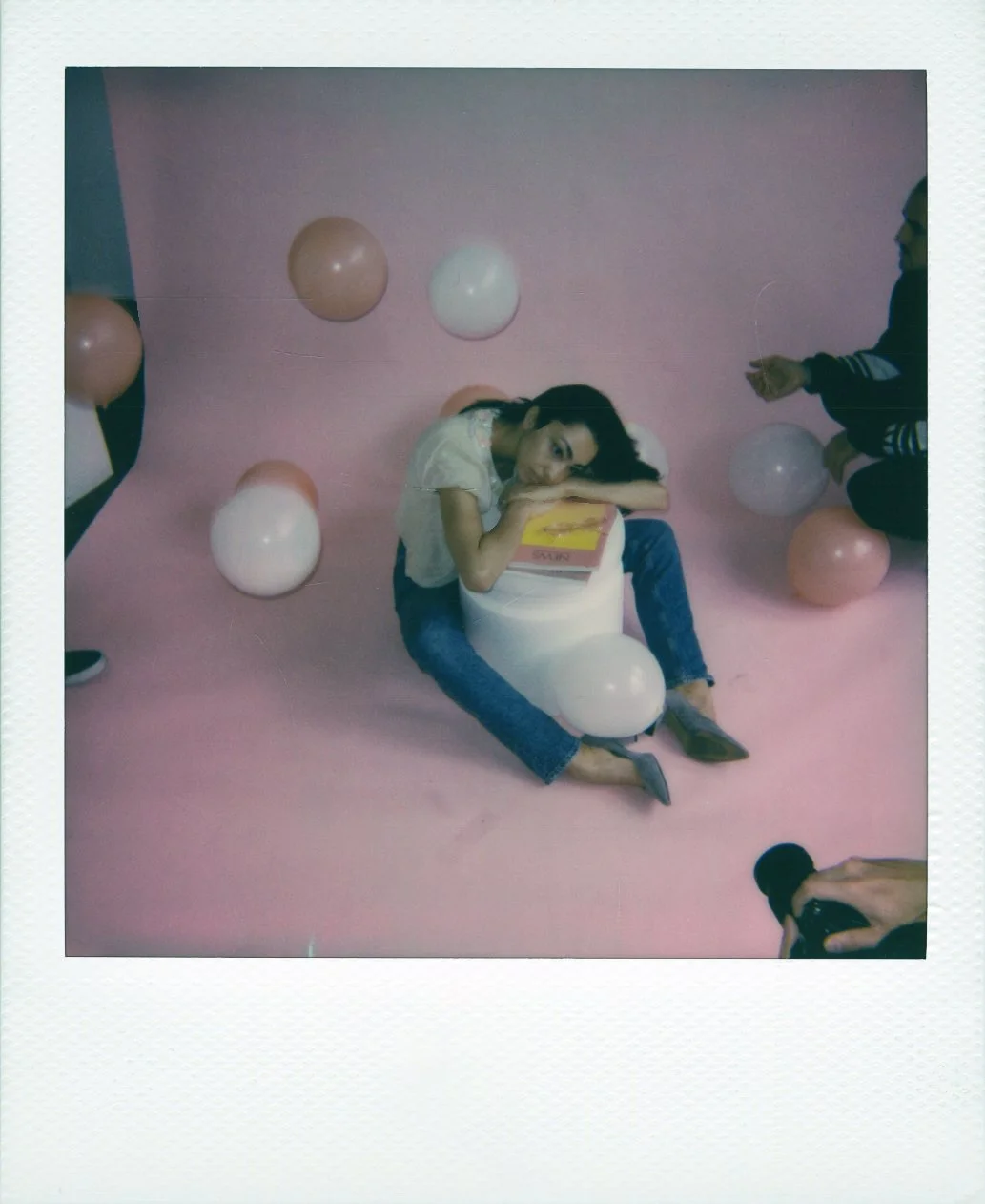 Square-frame polaroid of a woman sitting on a pink floor with pink, beige, and white balloons around her, resting her head on her arms on a white box, with people around her taking photographs.