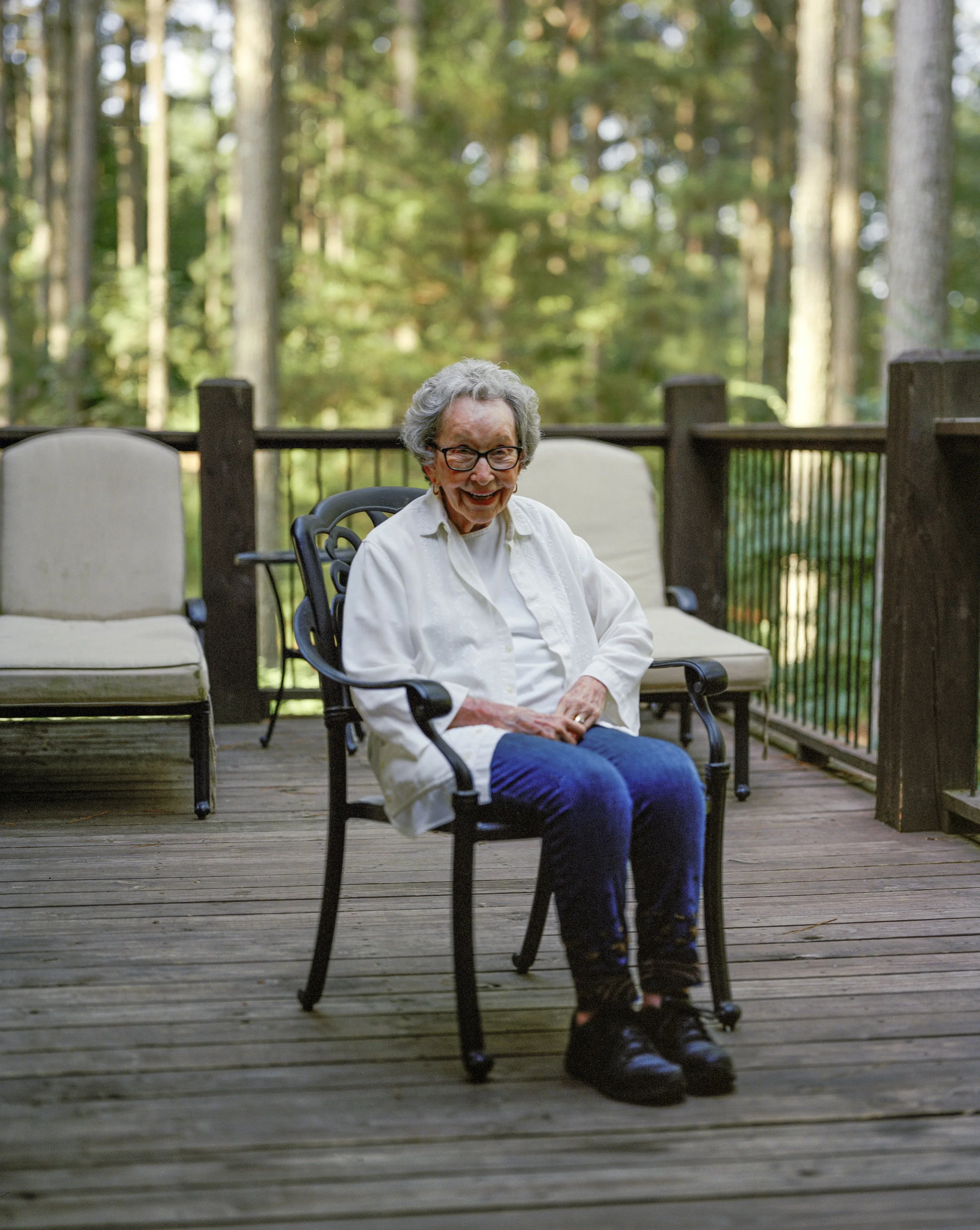 An elderly woman with glasses sitting on a patio chair outdoors, smiling, surrounded by trees.