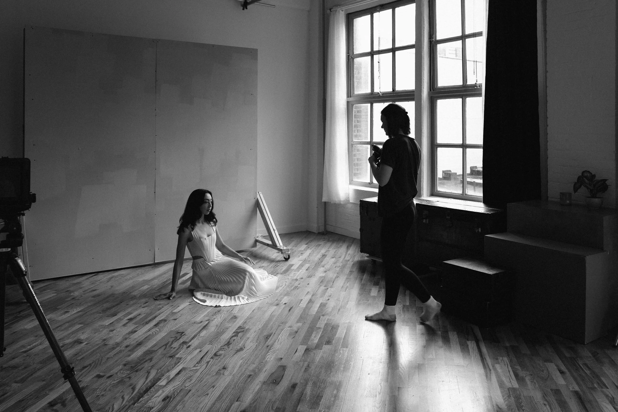 Black and white photo of a woman sitting on the wooden floor in a studio, with a photographer standing nearby. The woman is in a light-colored dress, sitting with legs crossed, facing the camera. The photographer is holding a camera, looking at the woman. Large windows provide natural light, and a large plain backdrop is behind the woman.