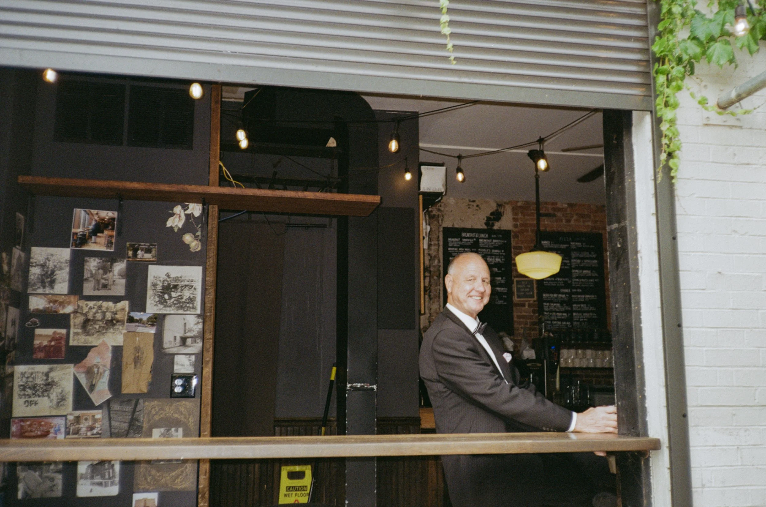 A smiling man in a black tuxedo with a bow tie stands behind a bar or counter in a venue with exposed brick walls, blackboard menus, and hanging light bulbs.