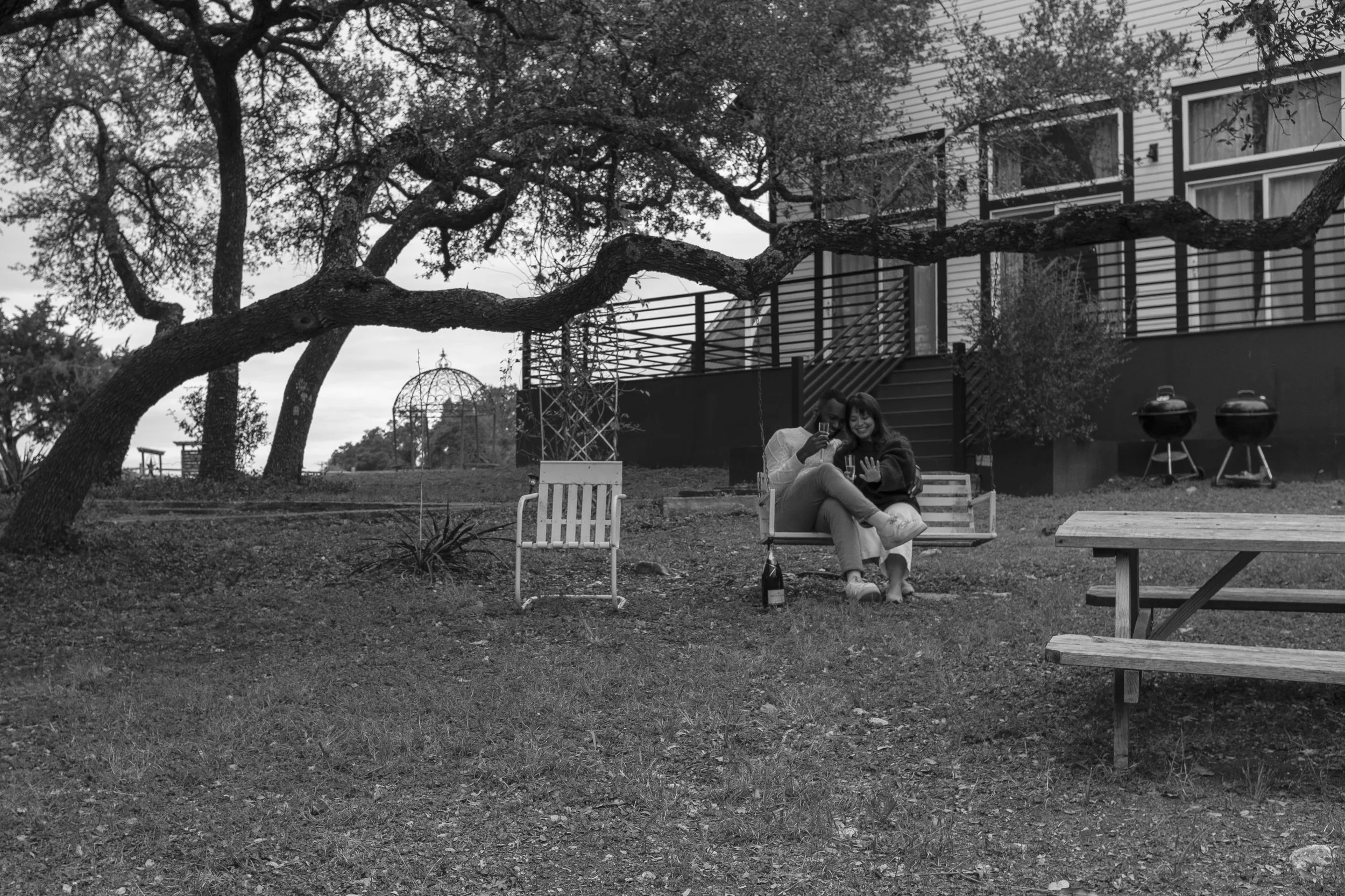 A couple sitting closely together on a bench in a backyard, admiring the woman's hand with a ring on it, surrounded by trees and outdoor furniture in black and white.