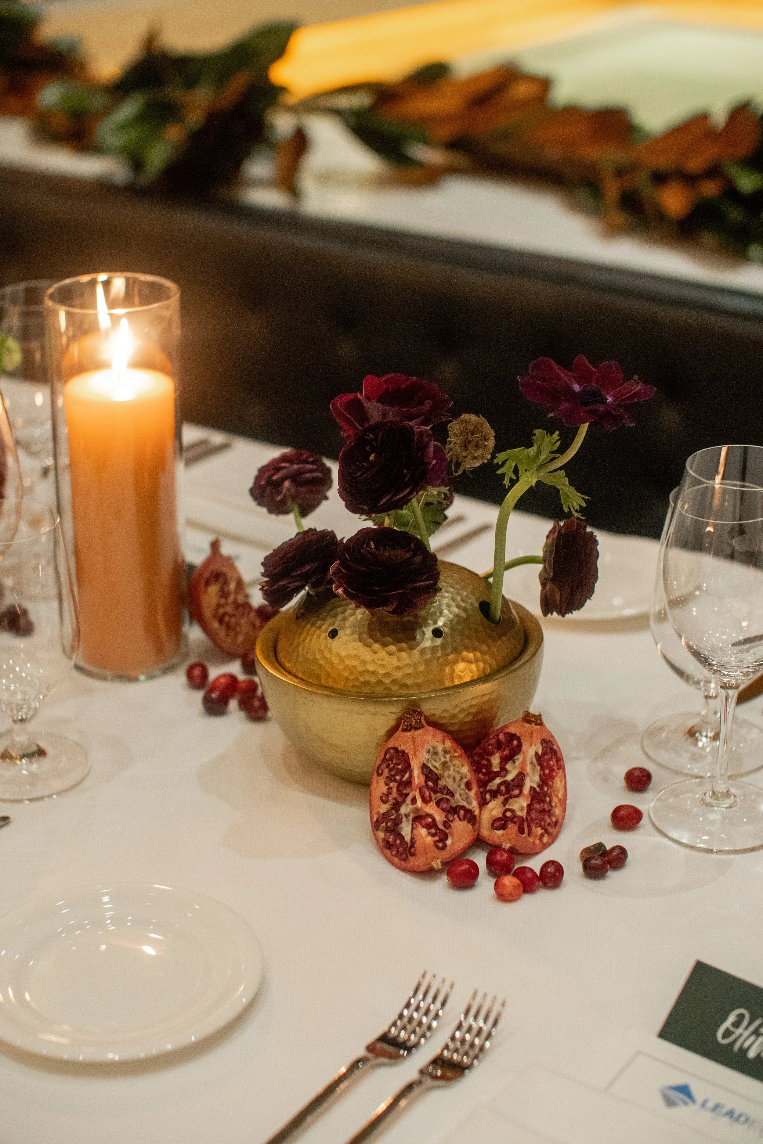 A table centerpiece with dark purple flowers in a hammered gold bowl, pomegranate halves, red berries, lit candle in a glass holder, wine glasses, and plates on a white tablecloth.