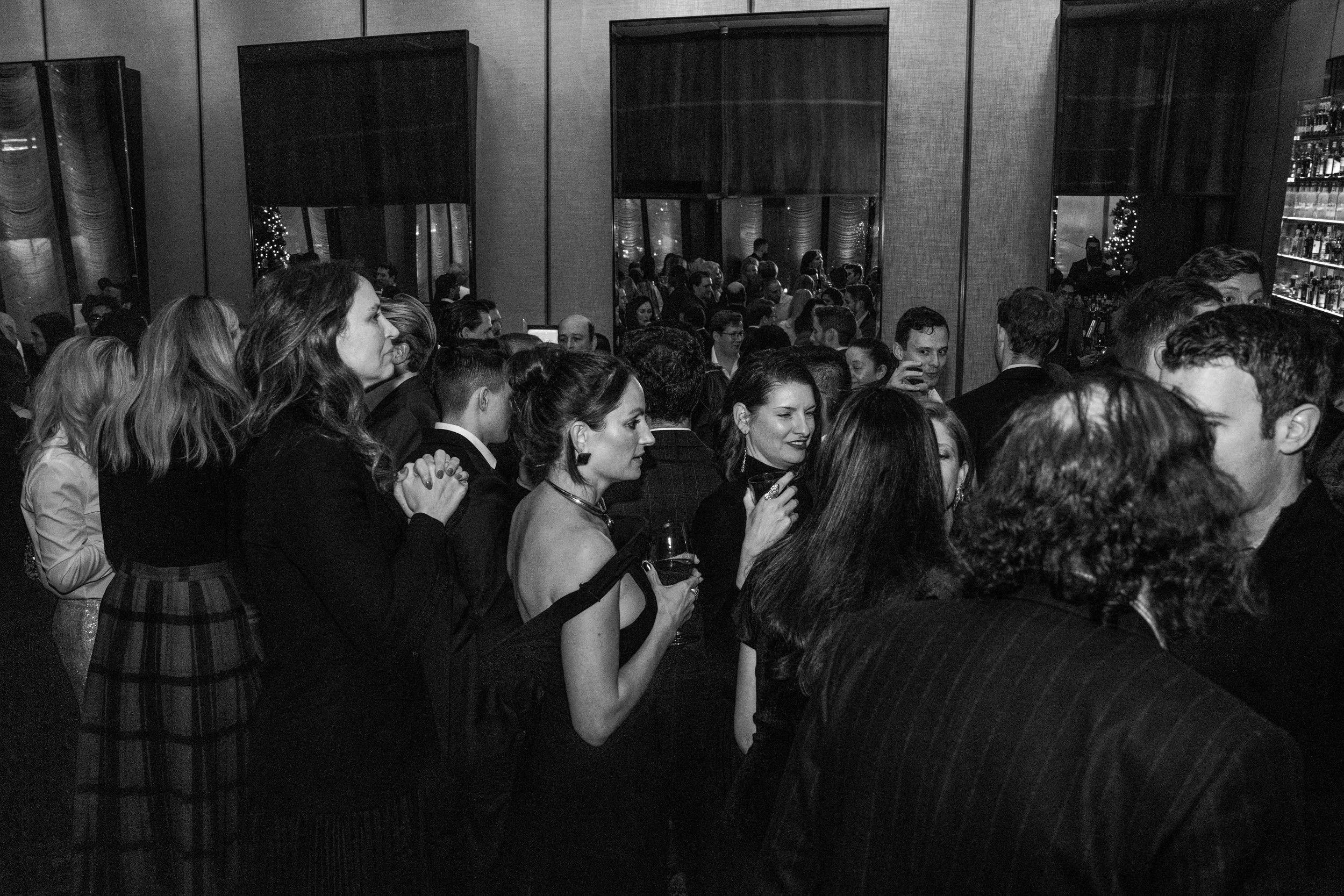 A crowded party or event with people standing, some holding drinks, in a dimly lit indoor space with mirrors and shelves in the background.