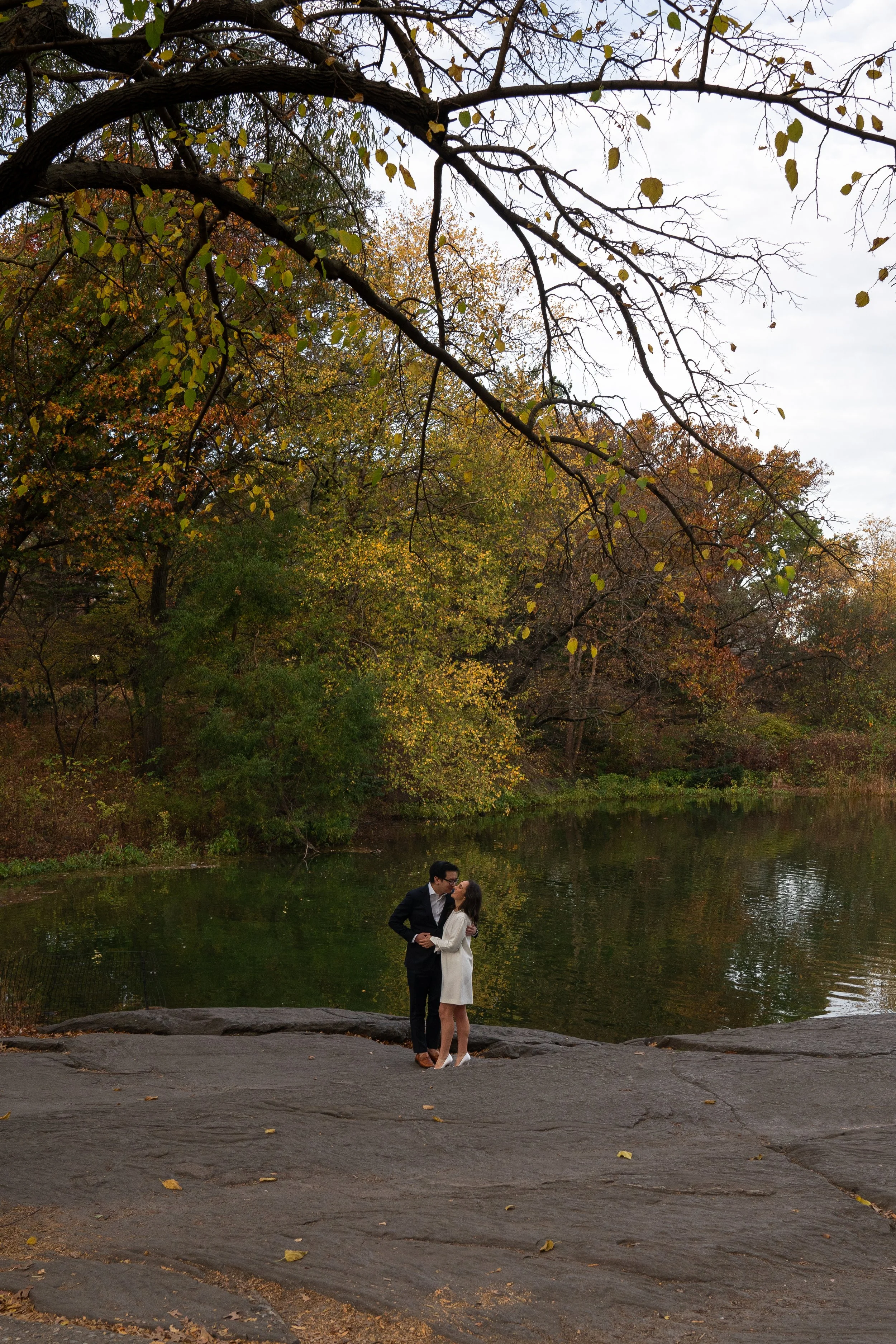 A couple in formal attire sharing a kiss by a lake during fall, surrounded by trees with yellow and green leaves.