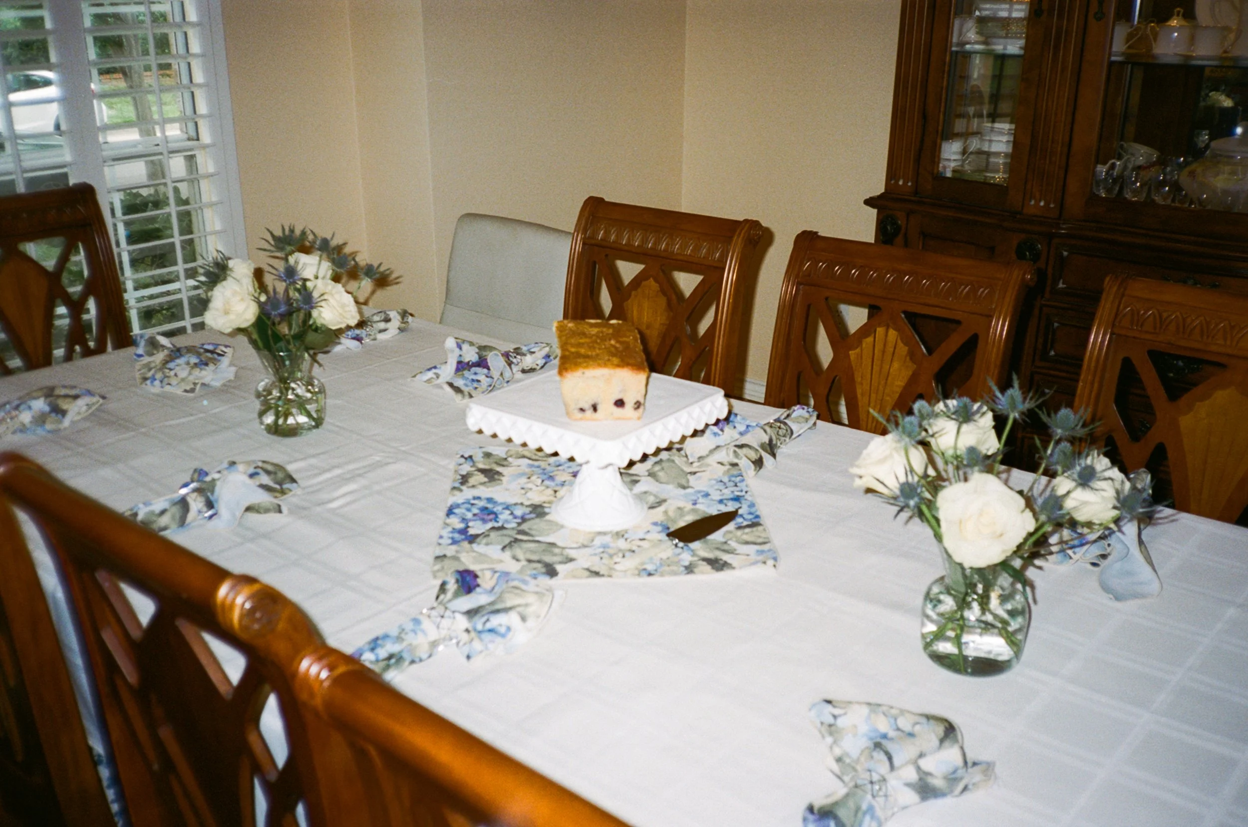 A dining table decorated with floral arrangements in vases, napkins, and a partially eaten cake on a cake stand.