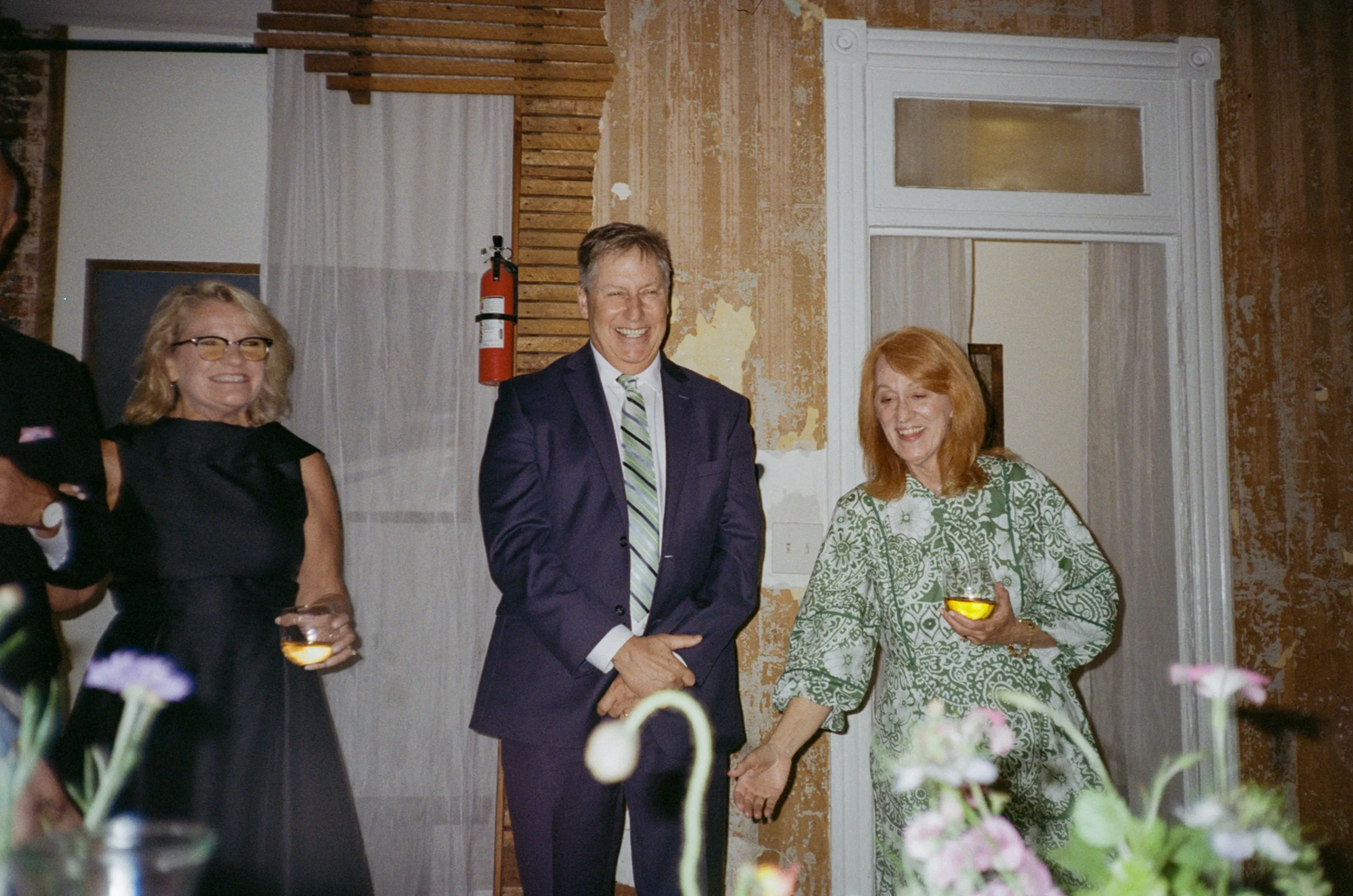 Three people standing indoors, smiling and holding glasses of yellow-colored drink, with a woman in black dress, a man in a navy suit, and a woman in green patterned dress.