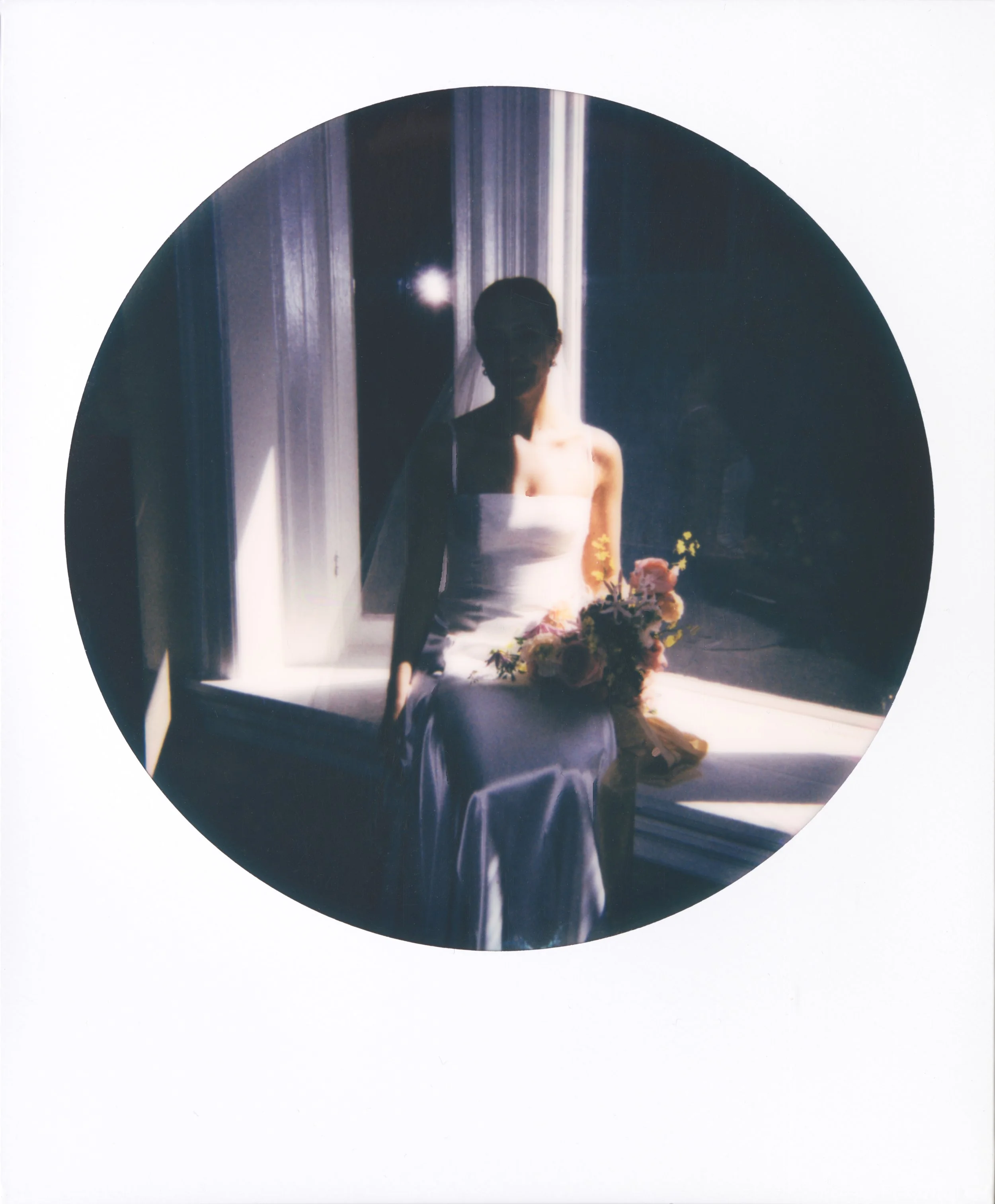Round-frame polaroid of a silhouette of a woman in a white dress holding a bouquet of flowers, sitting by a window with sunlight shining through.