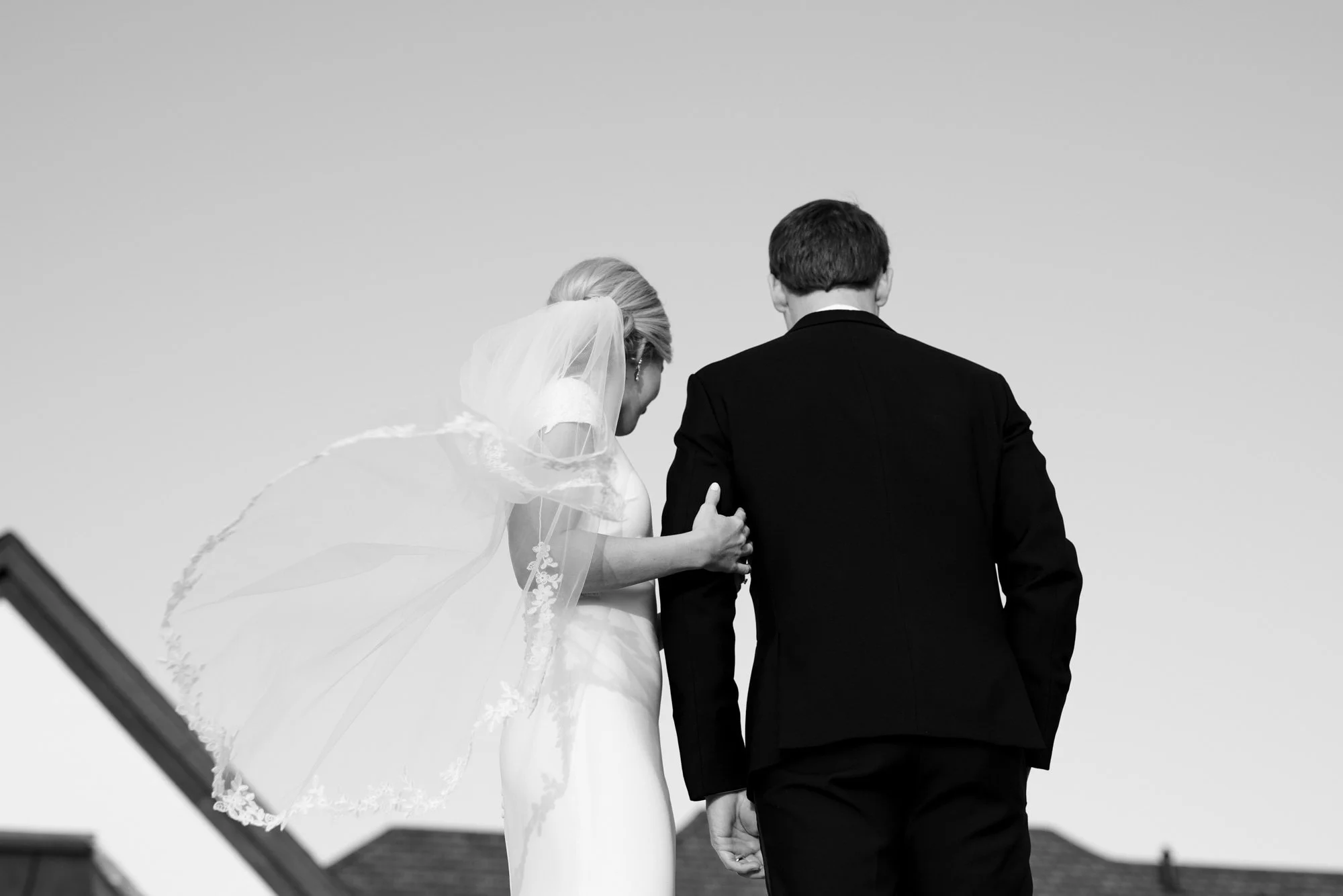 Black and white photo of a bride and groom holding hands, with the bride in a wedding dress and veil, outdoors.