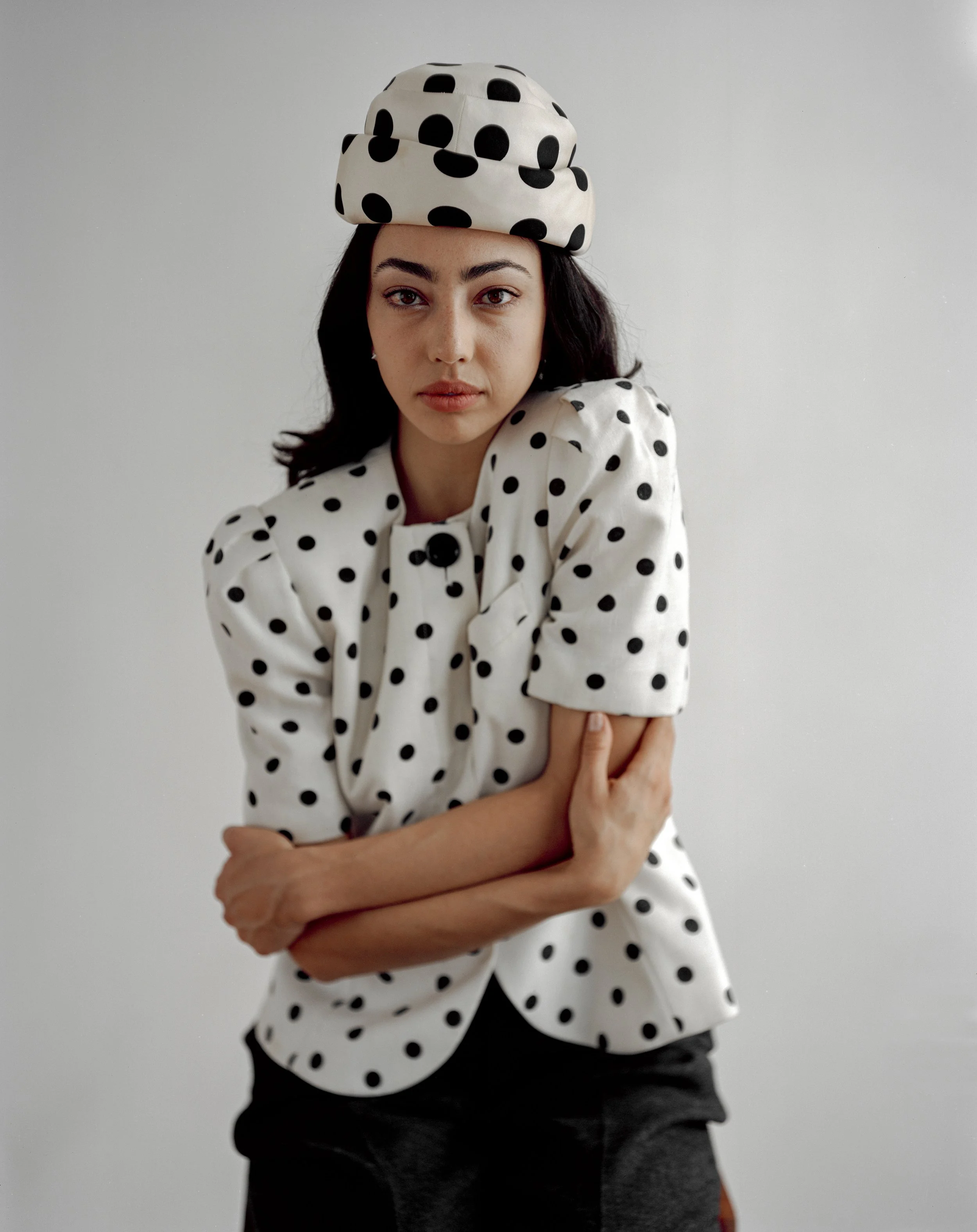 A woman wearing a black and white polka dot outfit, including a hat, shirt, and pants, posing with her arms crossed in front of a plain white background.