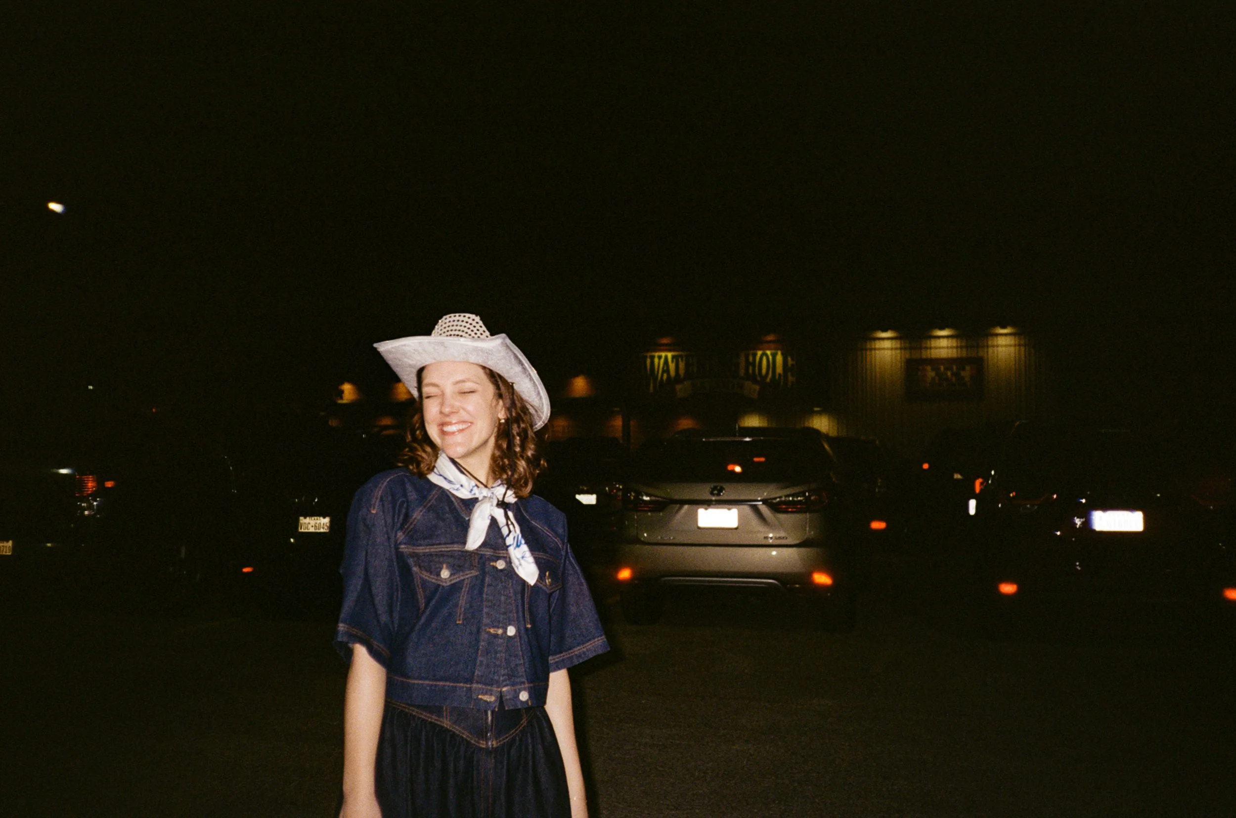 A smiling woman wearing a white wide-brimmed hat, denim jacket, and neck scarf, standing in front of a parking lot at night.