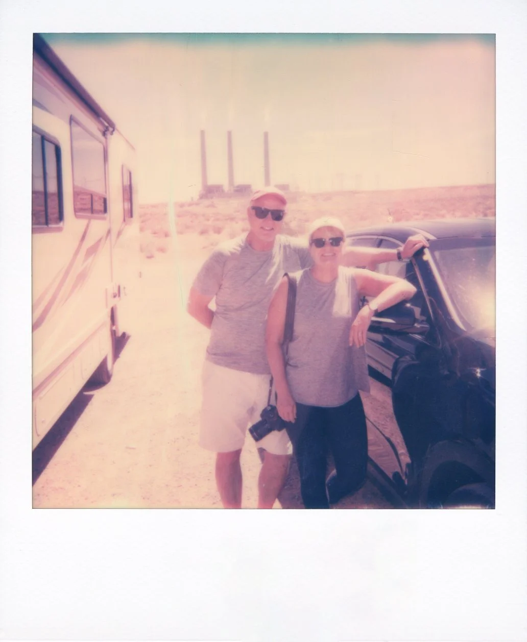 Square-frame polaroid of two people standing outdoors next to a black car and a yellow camper van, with power plant smokestacks in the background, bright sunny day, wearing sunglasses and casual summer clothing.