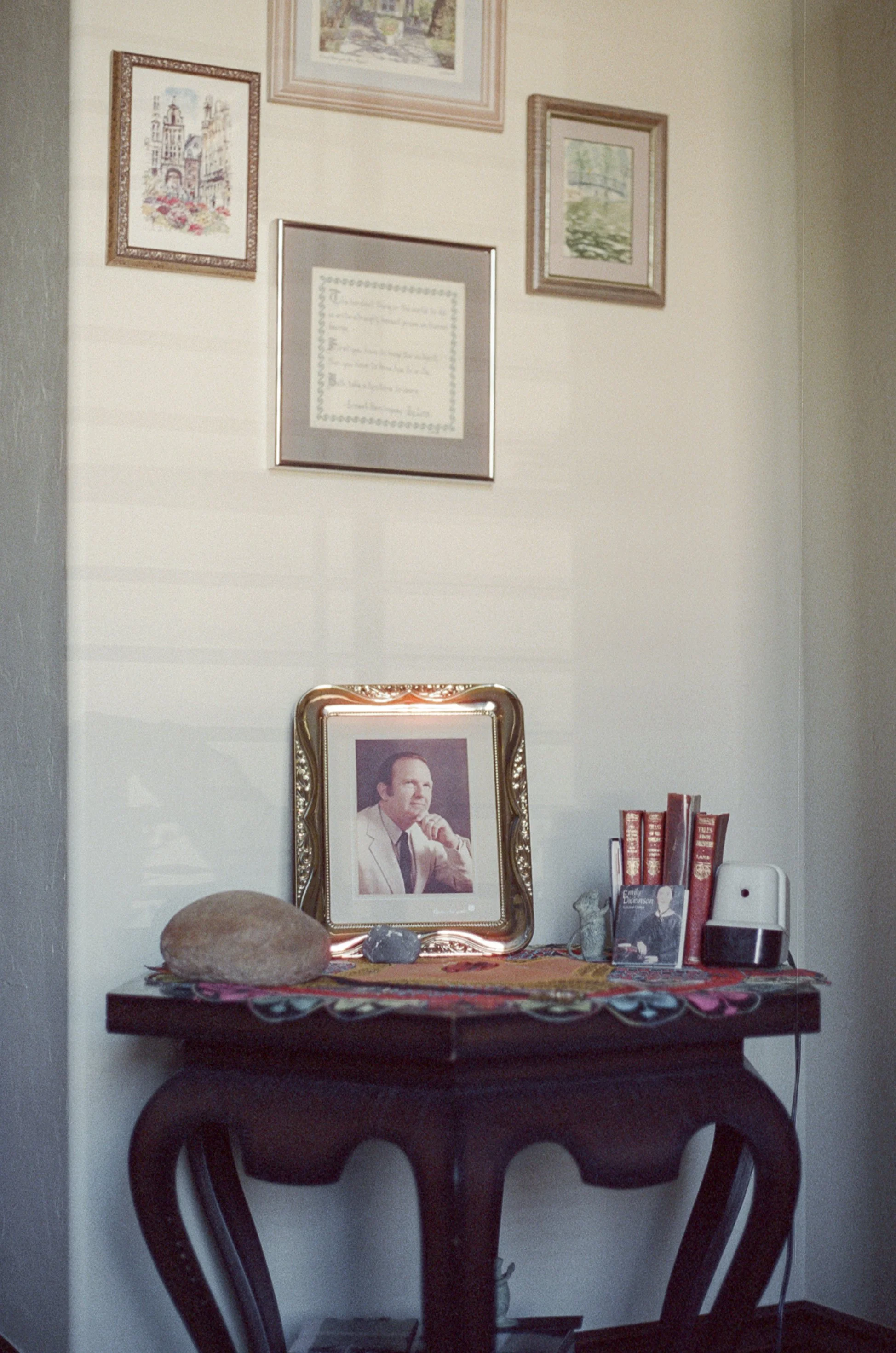 A framed photo of a man in a white suit on a decorative table. The table has rocks and small sculptures, books, and an essential oil diffuser. Above the table on the wall, there are five framed pictures and documents.
