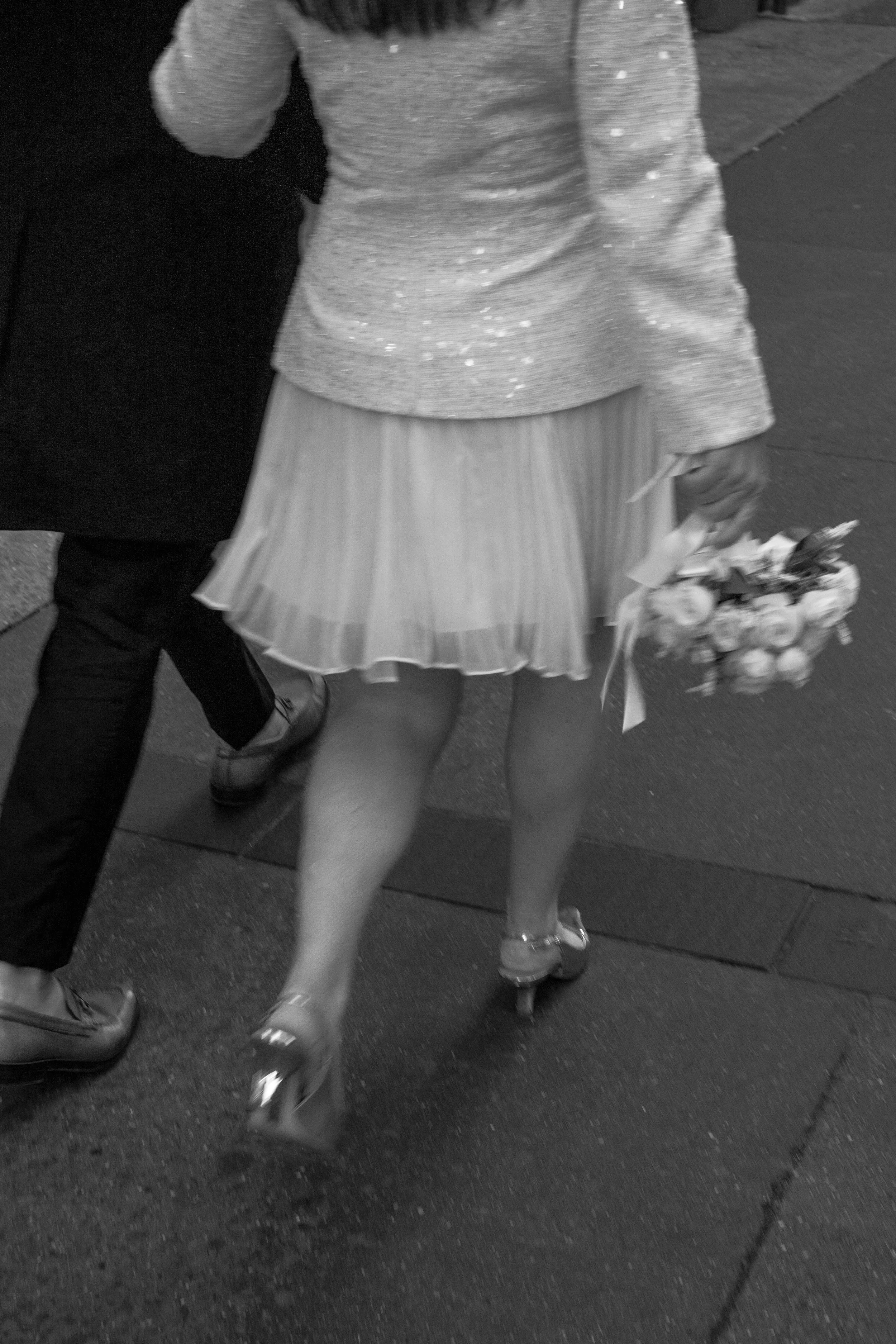 A woman wearing a glittery top, a pleated skirt, and high heels, walking with a bouquet of flowers in her right hand.