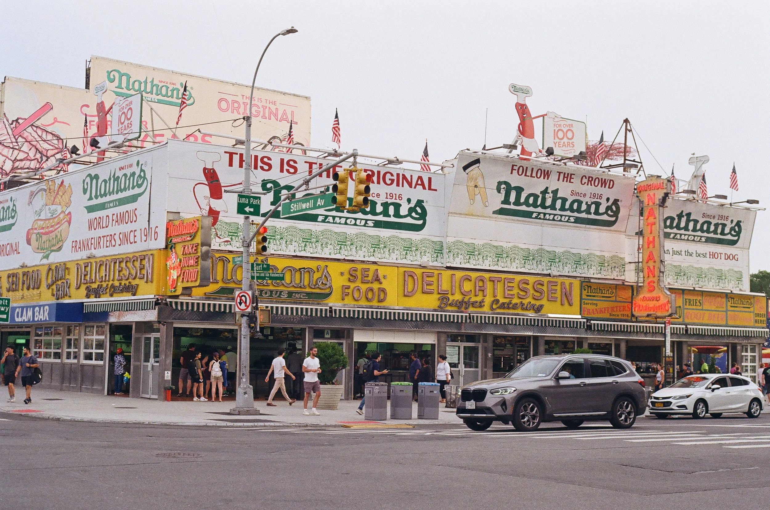 Nathan's Famous hot dog stand on a busy street corner, with pedestrians walking and cars parked in front. The building has bright signs advertising seafood, delicatessen, hot dogs, fries, and other fast food.