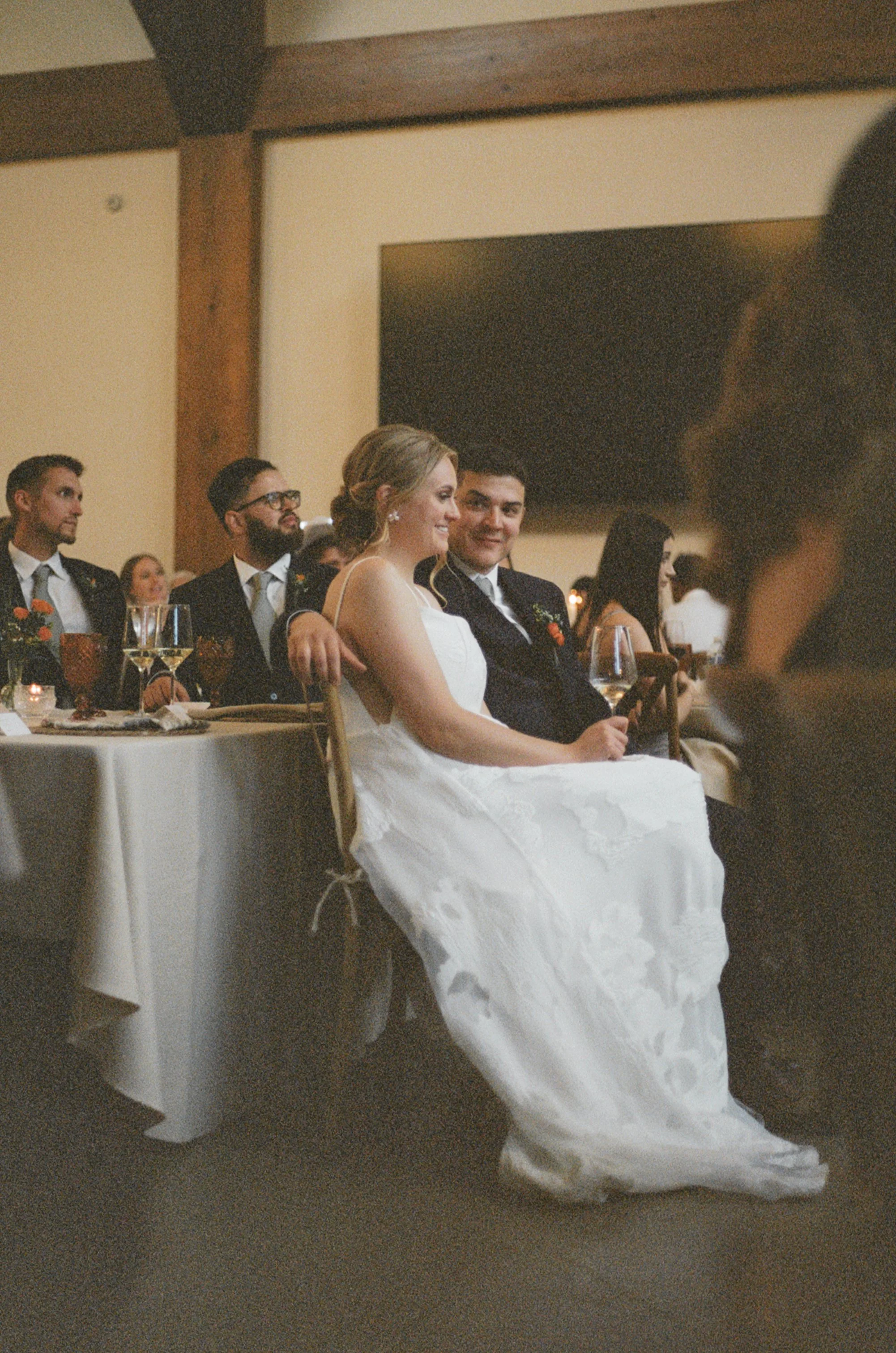 A bride and groom sitting at a reception table, surrounded by wedding guests.