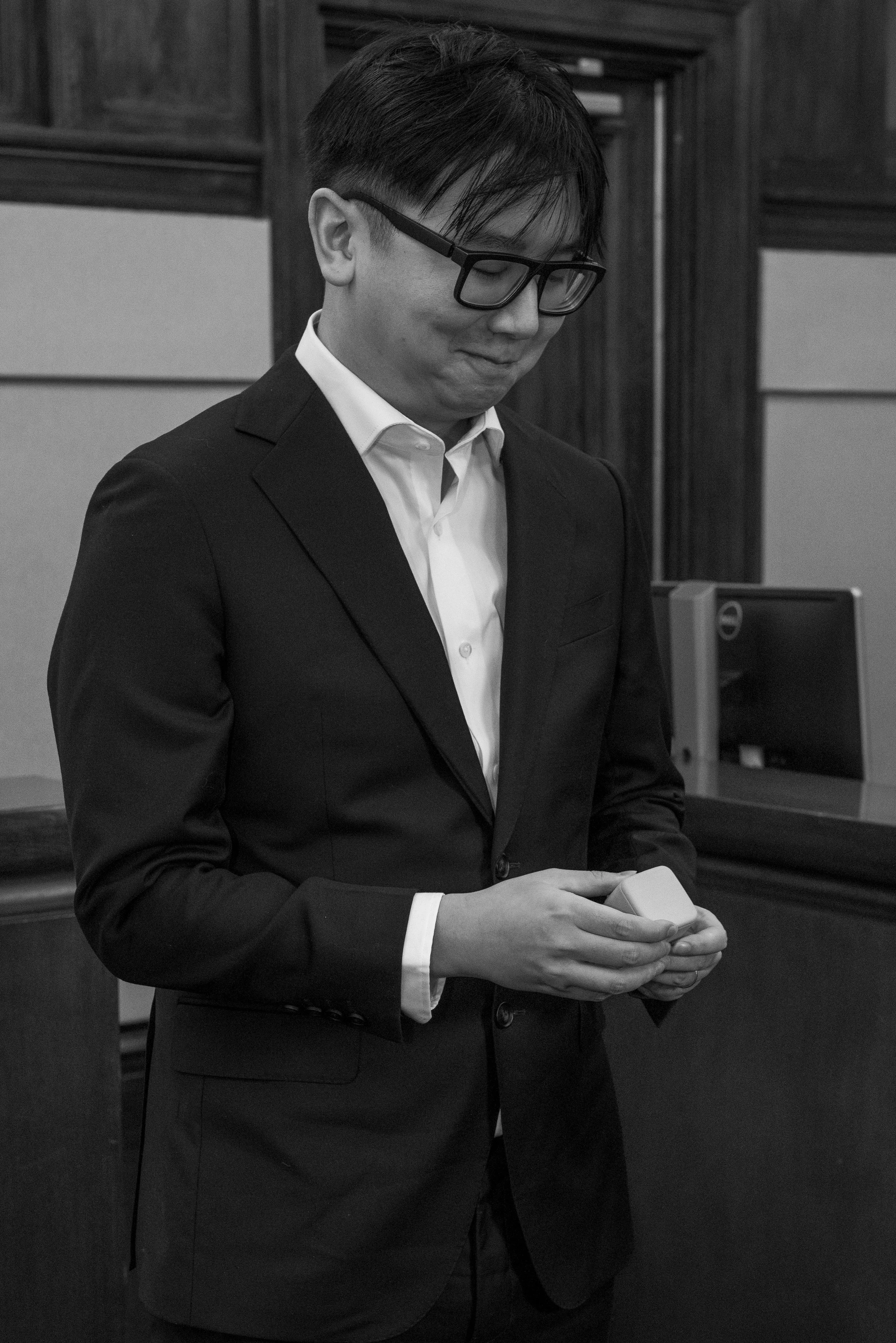 A man in a suit with glasses holding a small ring box,in his hands. He is indoors with wood paneling, likely a courtroom.