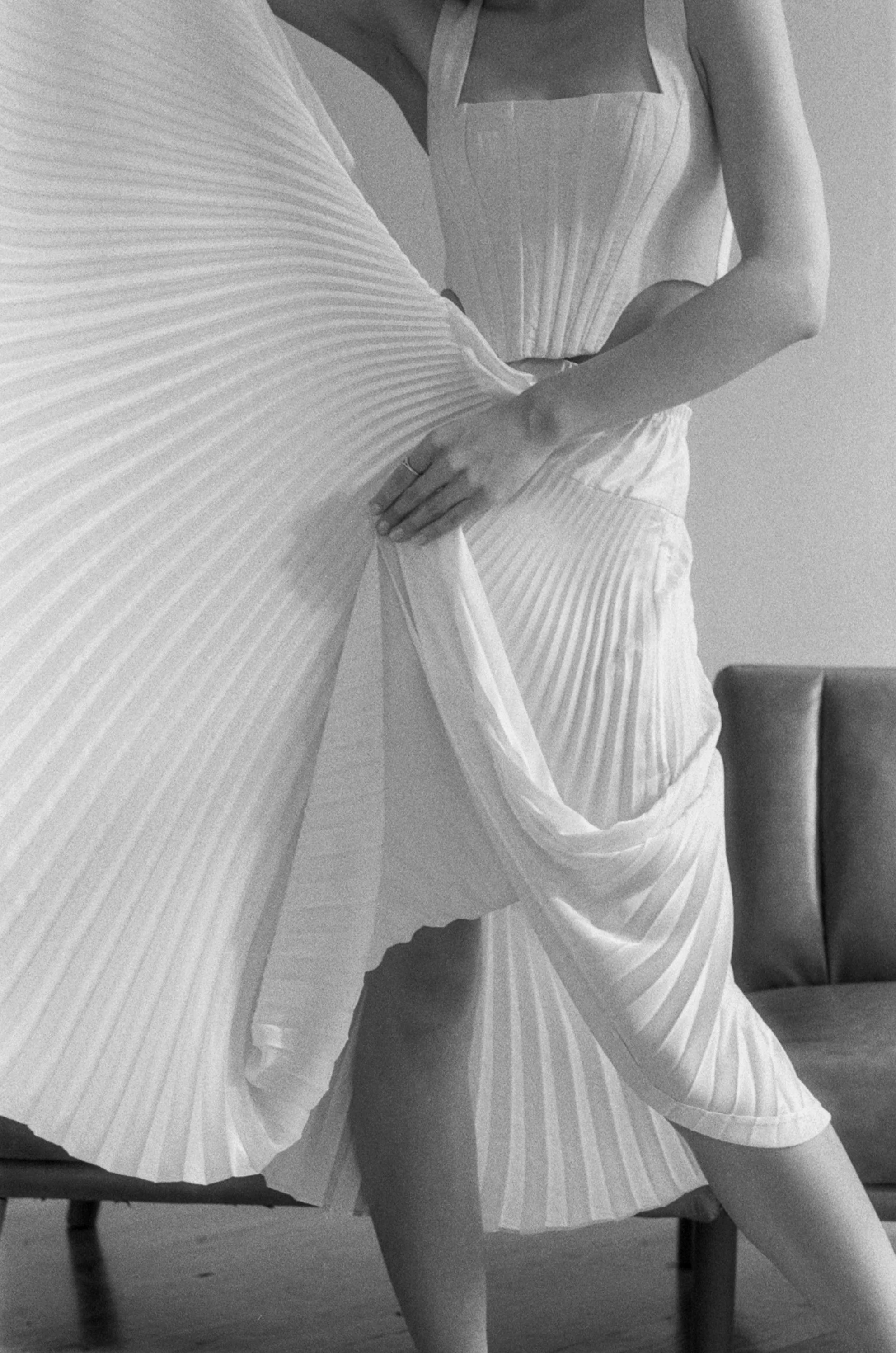 A person wearing a striped pleated dress, lifting the skirt to show the fabric's texture, with a gray sofa in the background.