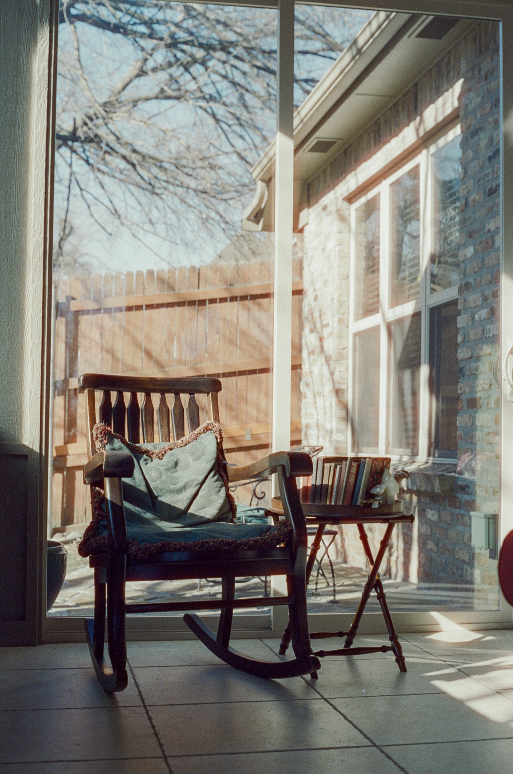 A wooden rocking chair with a cushion on it, placed on a tiled floor inside a house. Beside the chair, there is a small table with books and a decorative object. Behind the glass door, a backyard with a fence and brick house wall is visible, with leafless tree branches overhead and sunlight casting shadows.