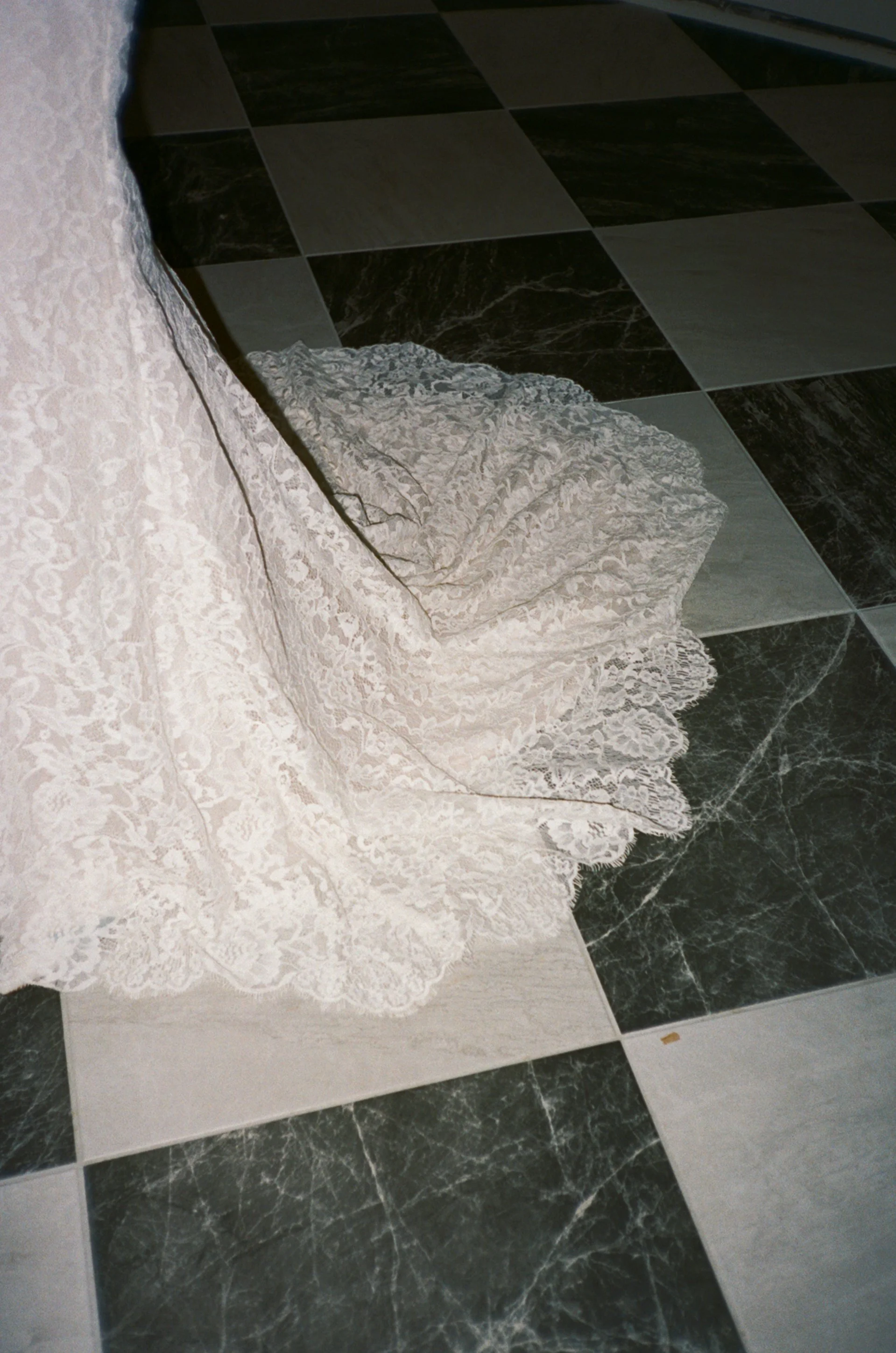 The train of a  white lace wedding dress spread out on a black and white marble tiled floor.