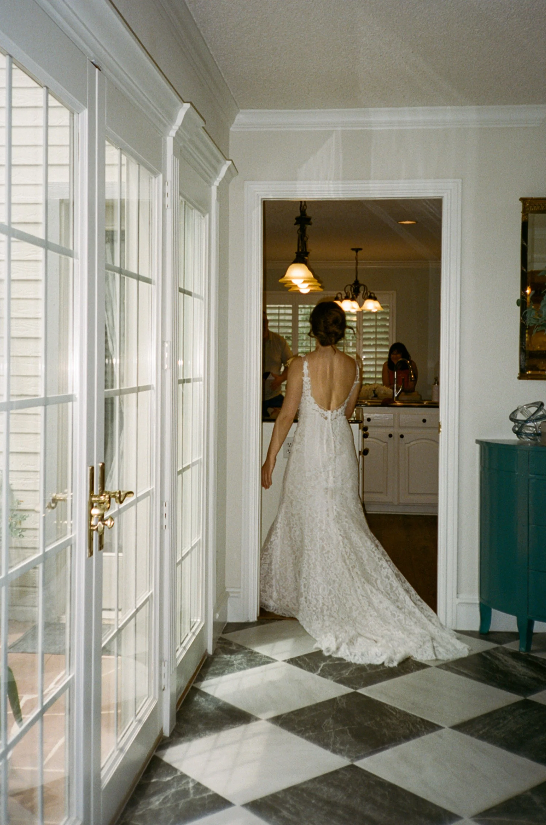 A bride in a white lace wedding dress walking into a room, seen from behind. The room has checkered black and white marble flooring and French doors to the outside.