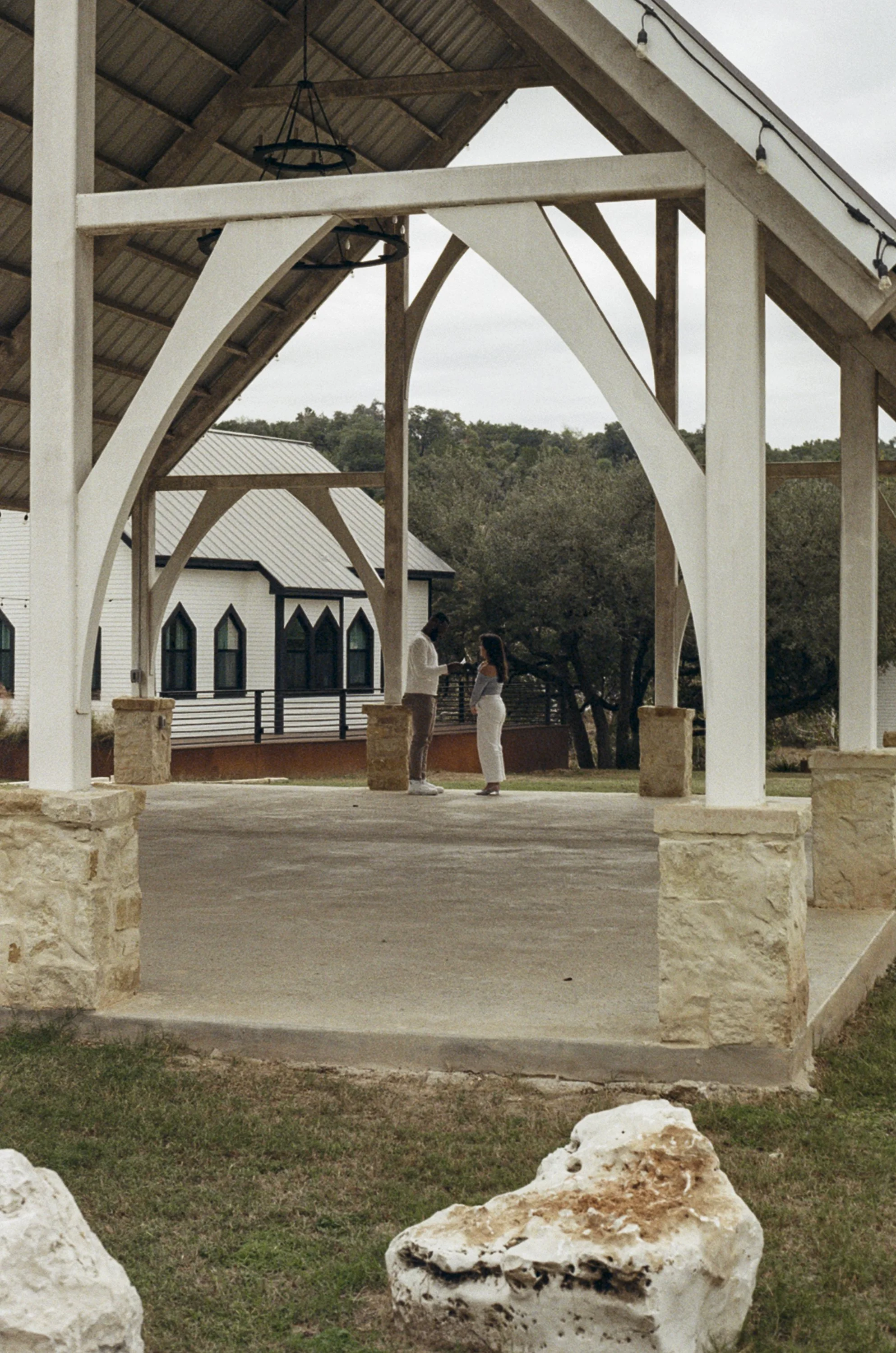 A man and woman standing under a wooden pavilion, engaged in conversation. The pavilion is supported by stone pillars, with a church building with pointed windows visible in the background, and trees surrounding the area.