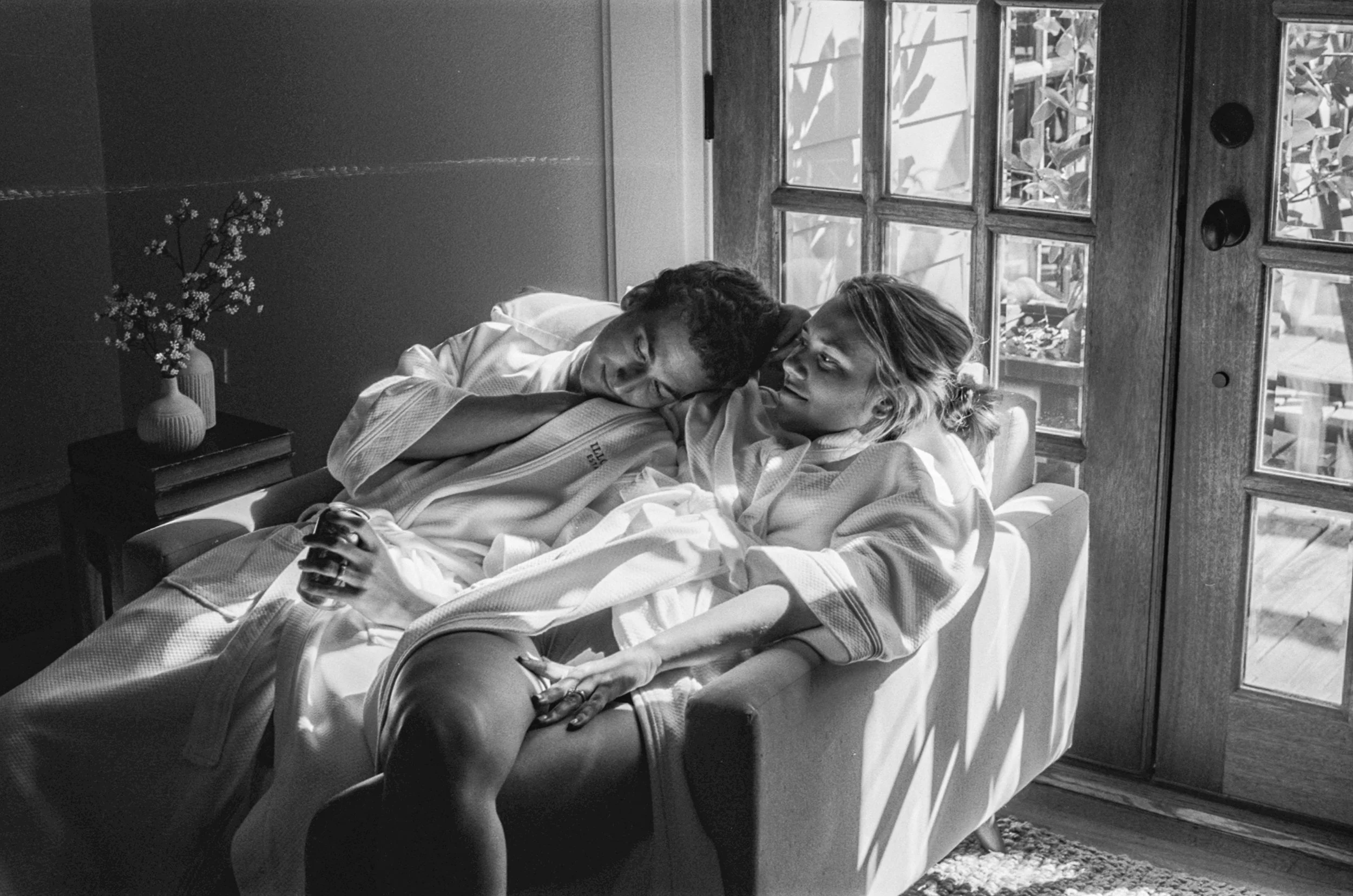 A black-and-white photograph of a man and woman lying on a couch in a cozy room with sunlight streaming through a glass door. The man is resting his head on the woman's shoulder while she looks at him affectionately, holding a glass in her hand. There is a small table with a vase of flowers and some books beside the couch.