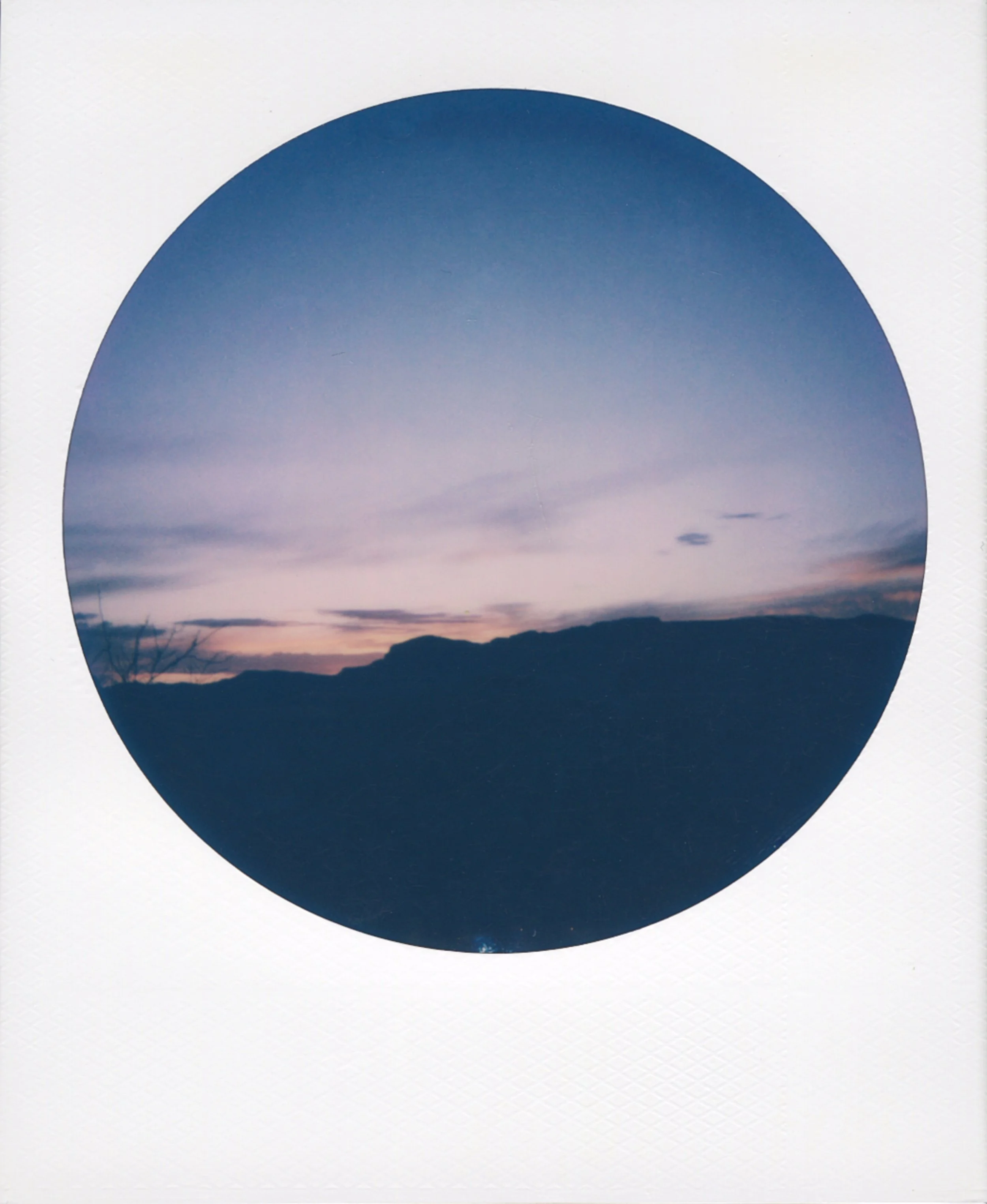 Round-frame polaroid picture of a sunset or dusk sky over dark silhouette mountains.