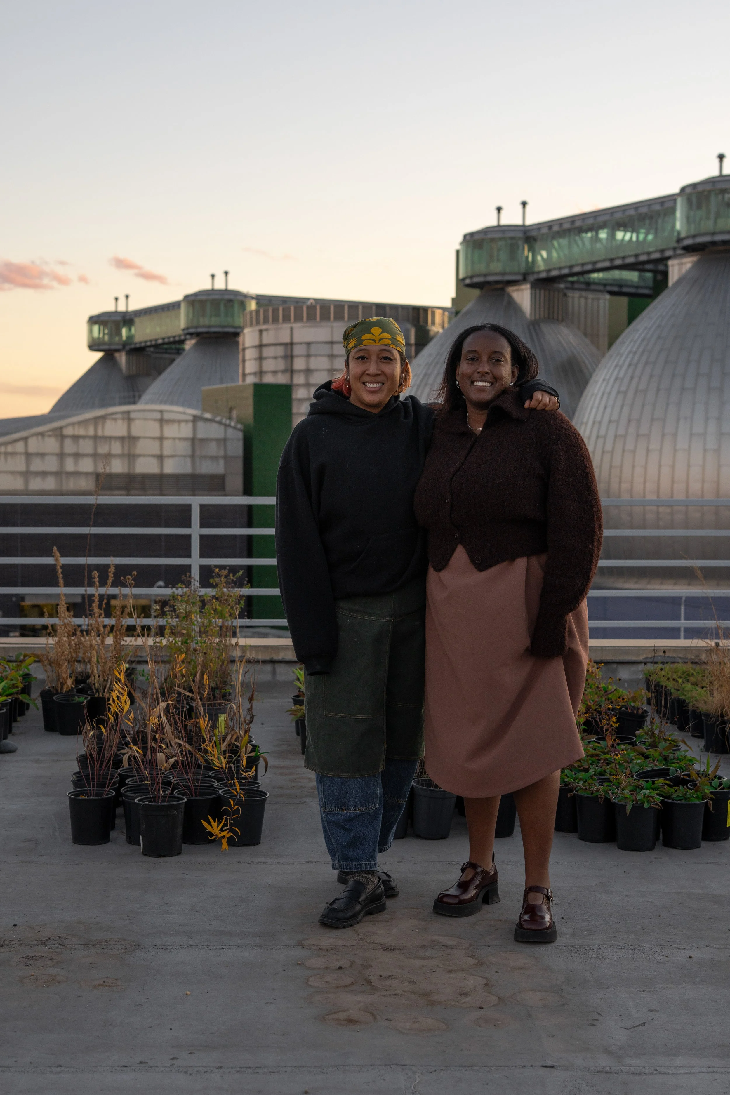 Two women standing close together on a rooftop during sunset, smiling at the camera. The background features large industrial or greenhouse structures, and potted plants are visible at their feet.