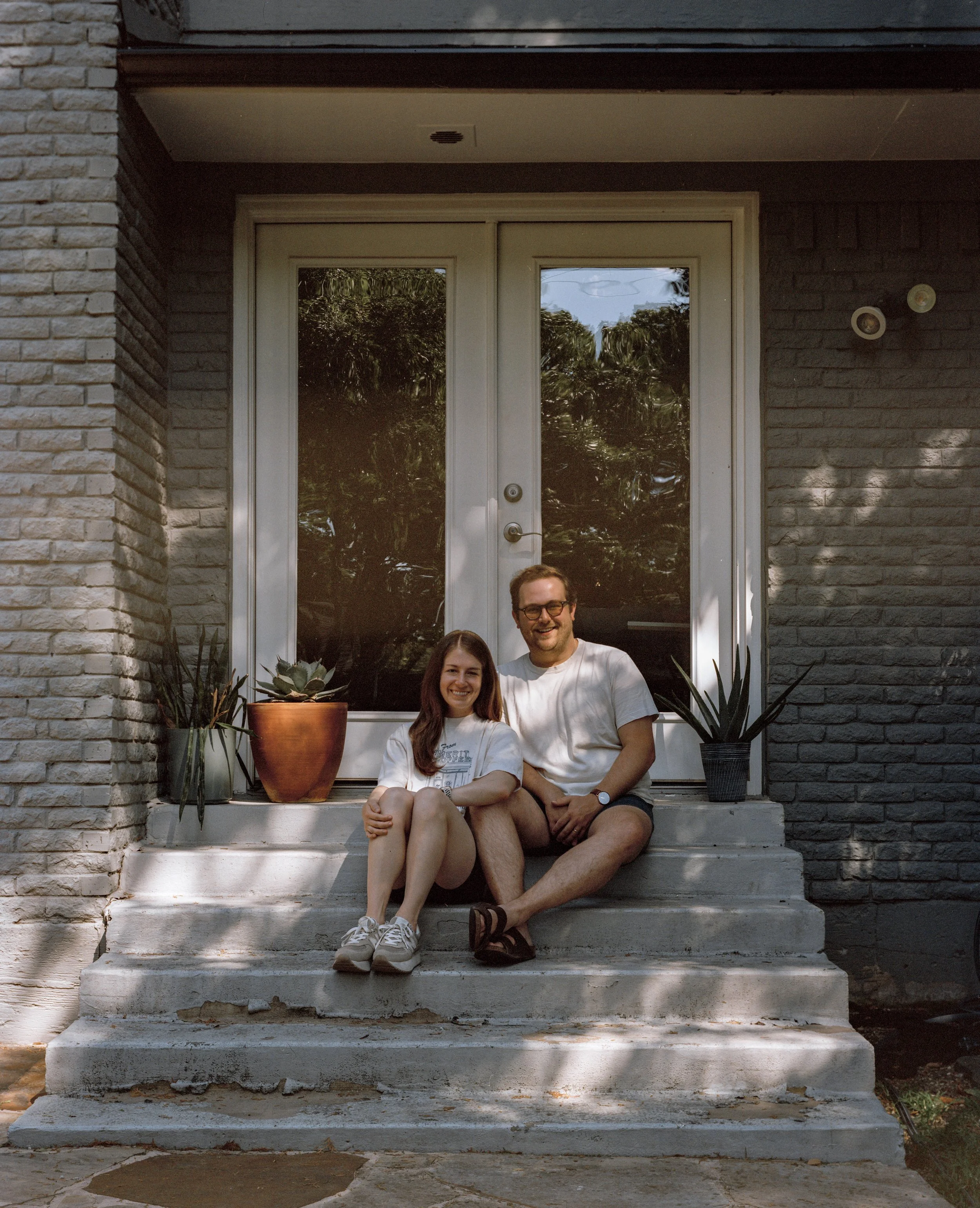 A smiling couple sitting on concrete steps in front of a glass door with potted plants on either side, outside a brick building.