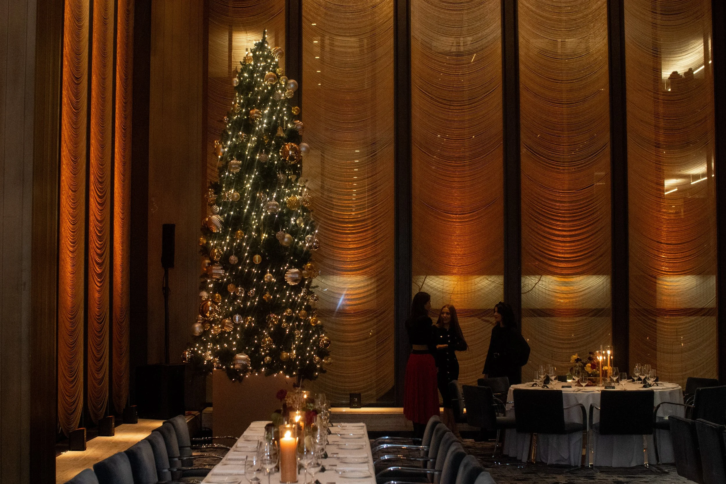 An indoor scene with a decorated Christmas tree with lights and gold ornaments, three women standing and talking near the window, and a table set for a celebration with candles and flowers.