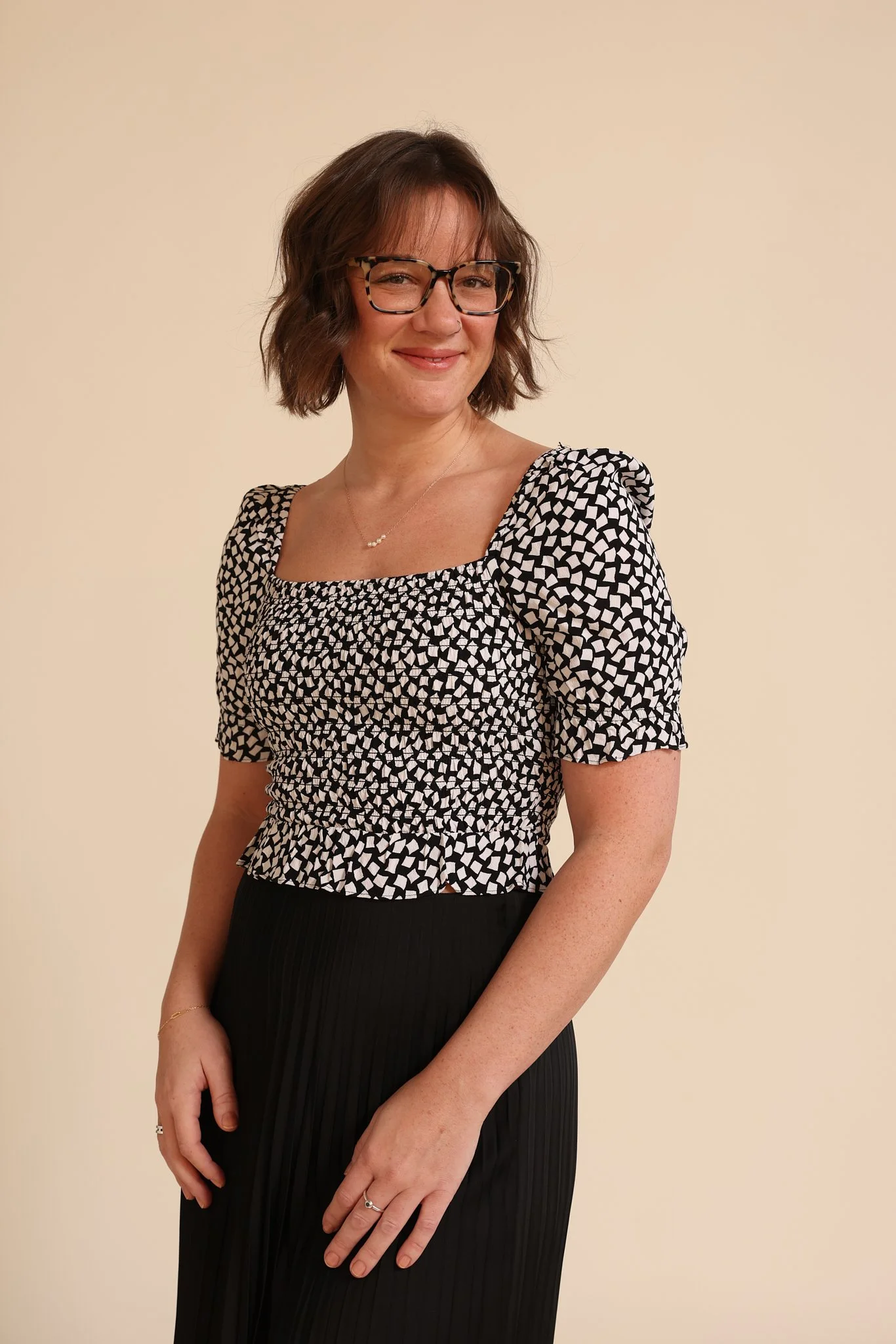A woman with short brown hair, wearing glasses, a patterned black and white top with puffed sleeves, and a black pleated skirt, smiling slightly, standing against a plain beige wall.