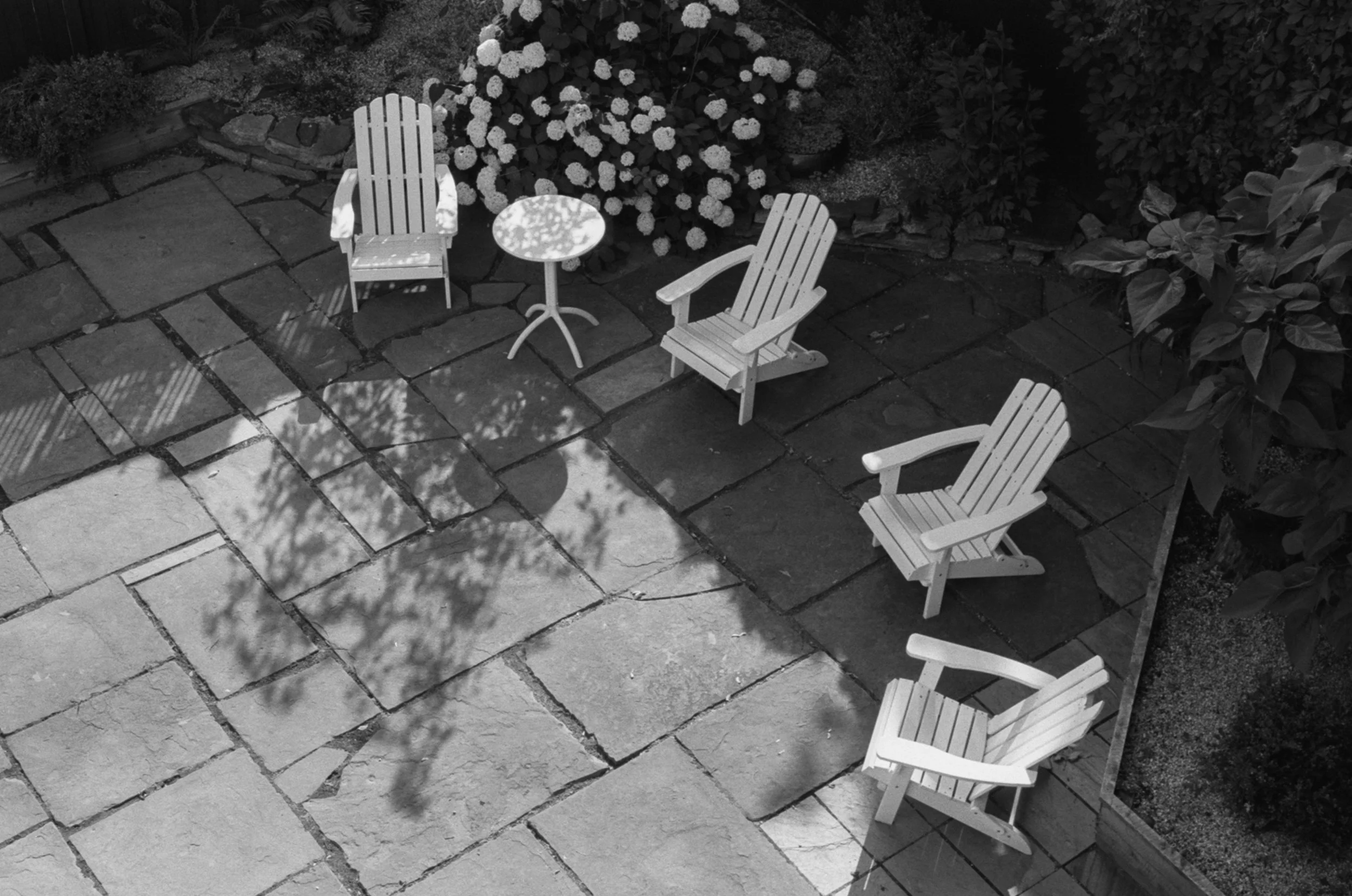 An outdoor patio with four wooden chairs and a small round table, surrounded by a garden with blooming flowers and plants, with sunlight casting shadows on the paved stone ground.