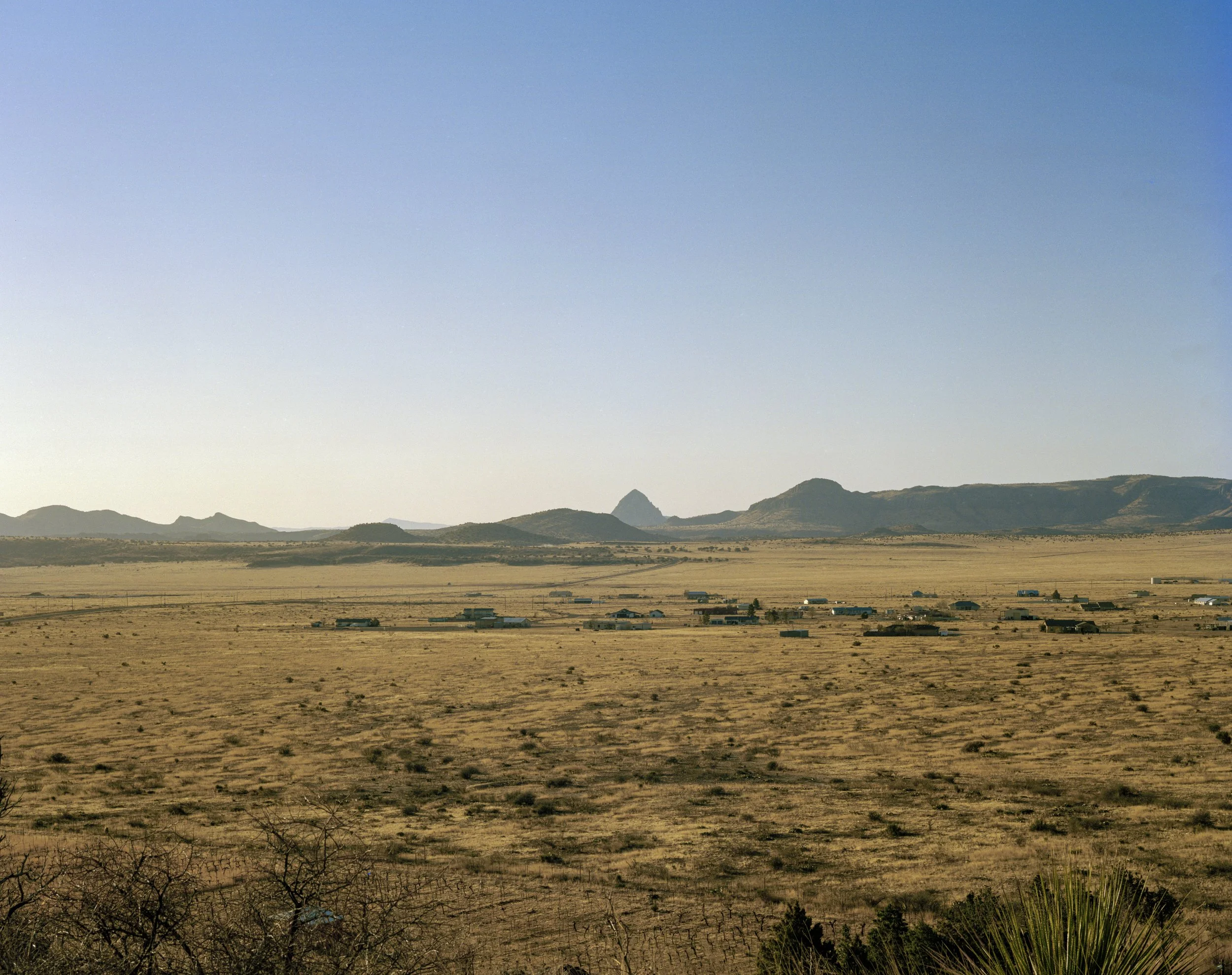 A vast desert landscape with scattered small buildings and distant mountain ranges under a clear blue sky.