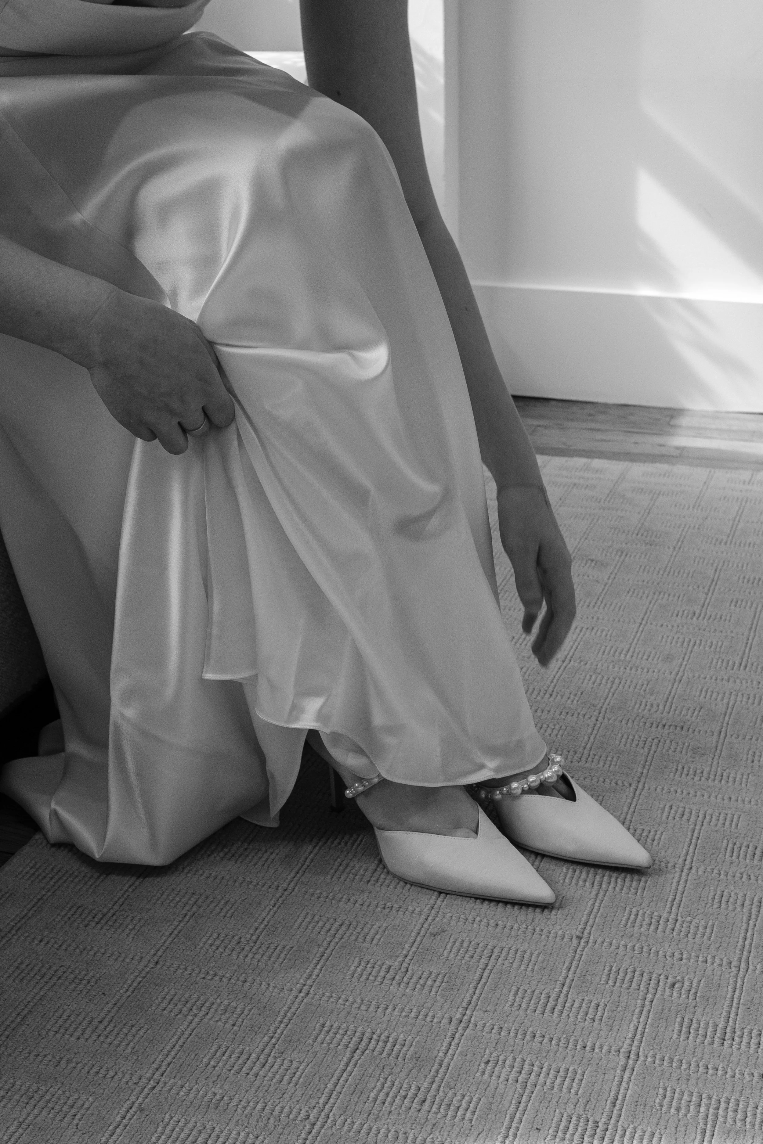 Close-up of a person wearing a satin wedding dress, and strapping on pointed-toe shoes.