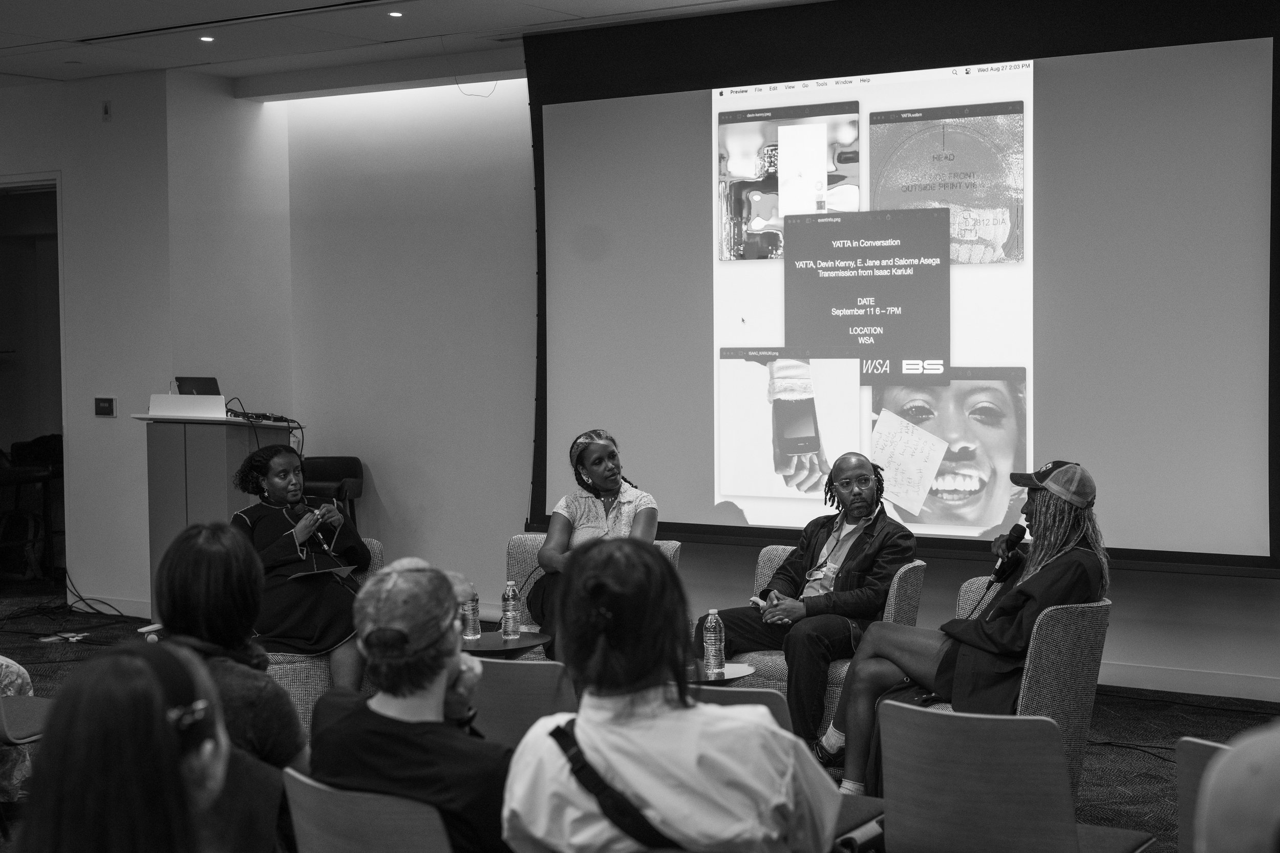 Panel discussion with four people seated on a stage in front of a large screen, audience watching. The large screen displays a digital presentation titled 'YATTA in Conversation,' with event details and images.
