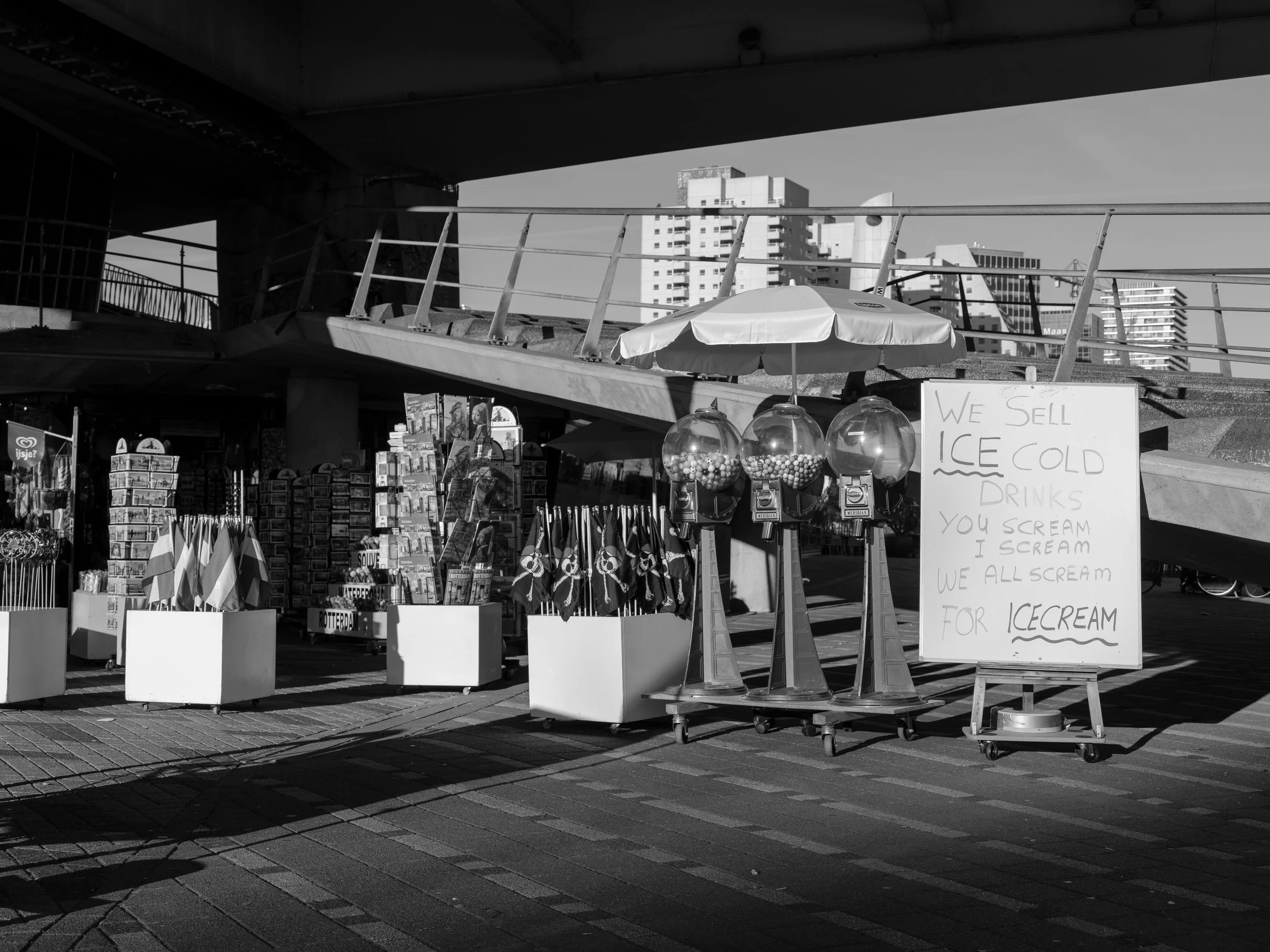 A street vendor stand selling ice-cold drinks and ice cream, with a handwritten sign, under an overpass in an urban area with tall buildings in the background.