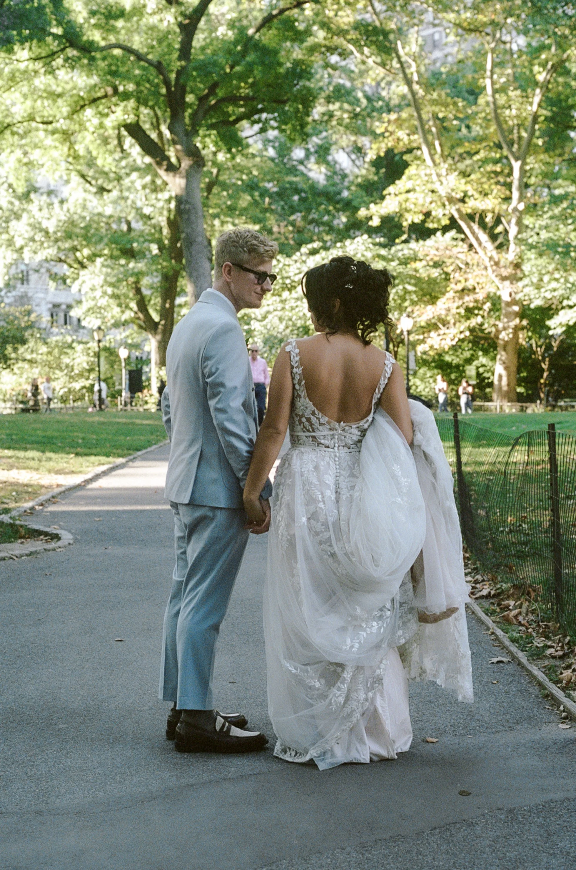 A newly married couple holds hands in a park, with the woman carrying her wedding dress over her arm. The man wears a light gray suit, and the woman wears a white lace wedding gown. They stand on a paved path surrounded by green trees.