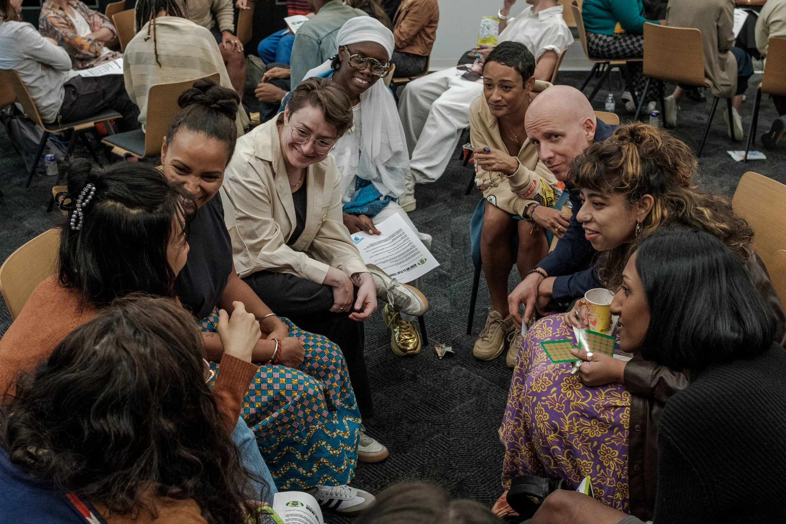 A diverse group of people sitting on chairs and in a circle, engaging in a discussion or activity at an indoor event.