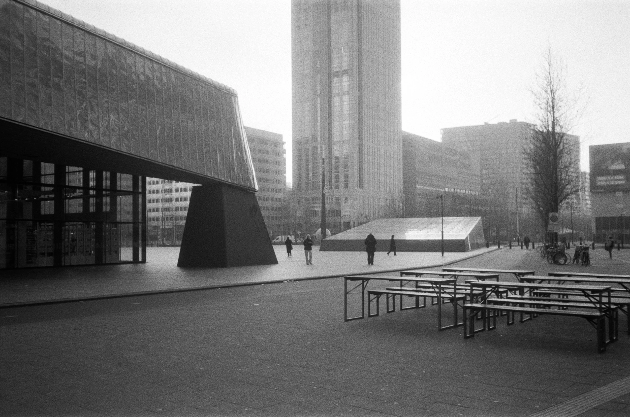 Black and white photo of a city square with modern buildings, people walking, bicycles, and metal tables in the foreground.