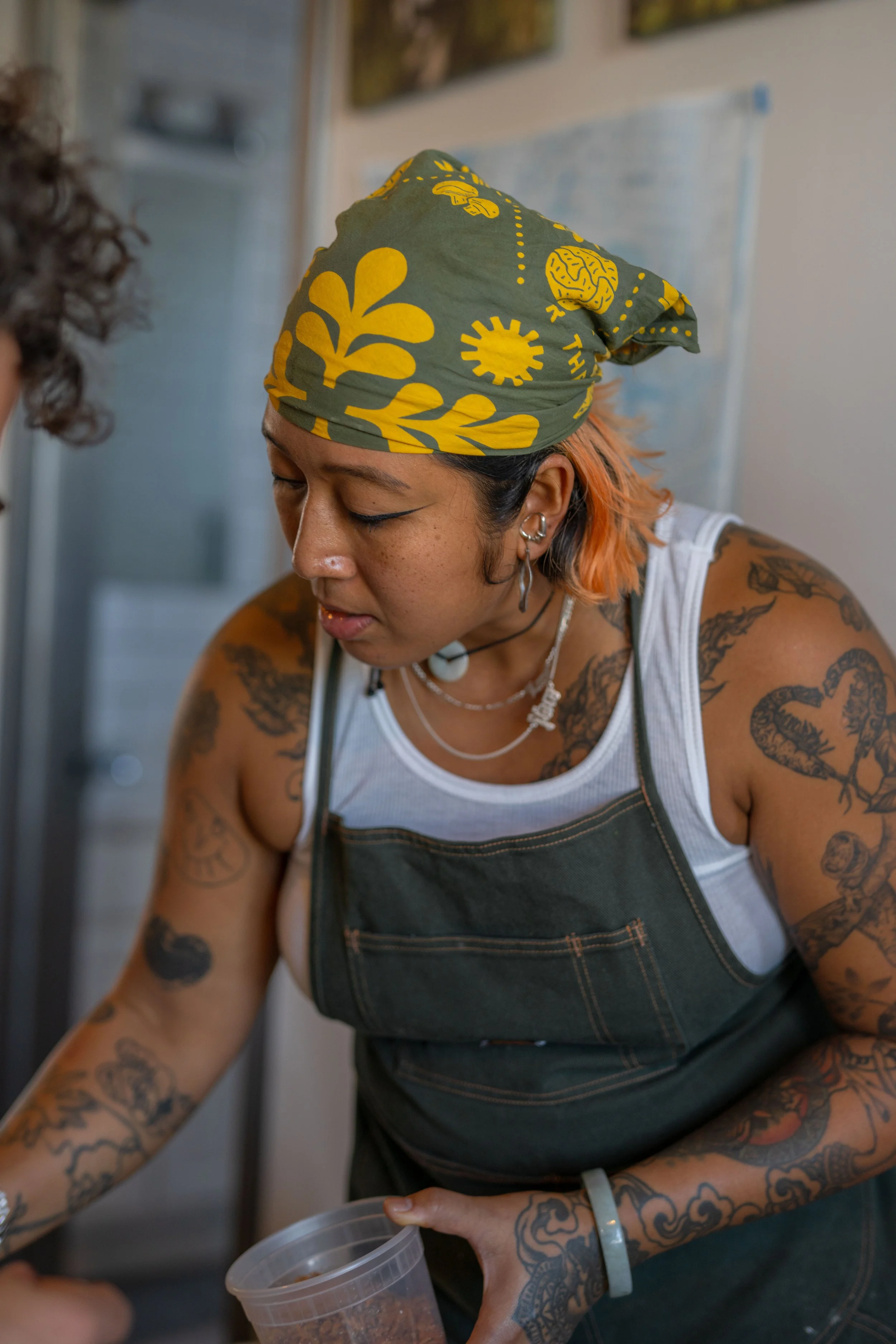 A woman with tattoos, wearing a green bandana with yellow patterns, a white tank top, and a dark apron, working in a kitchen.