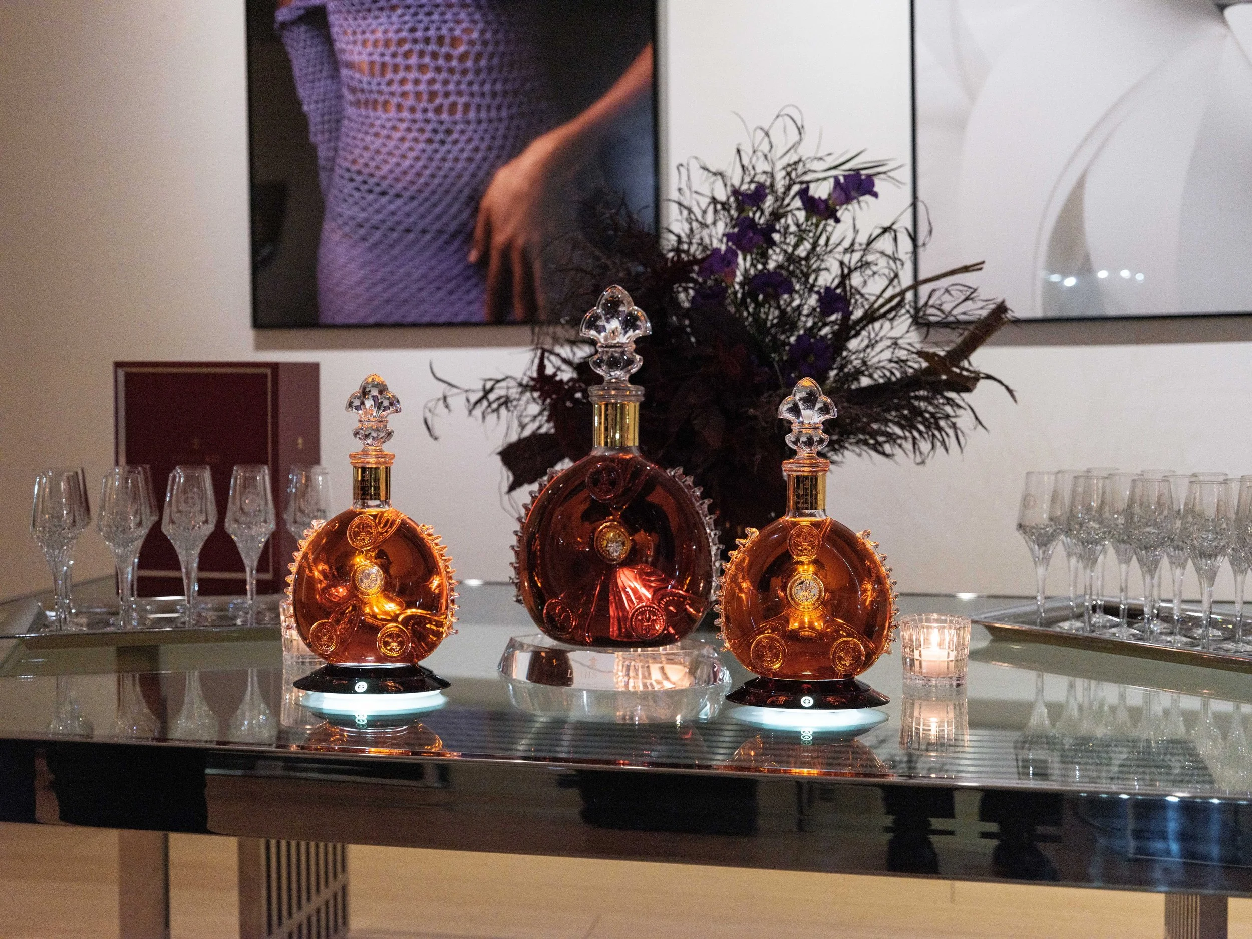 A table displaying three decorative bottles, a tray of empty glasses on each side, and a flower arrangement in the background, with a large portrait on the wall behind.